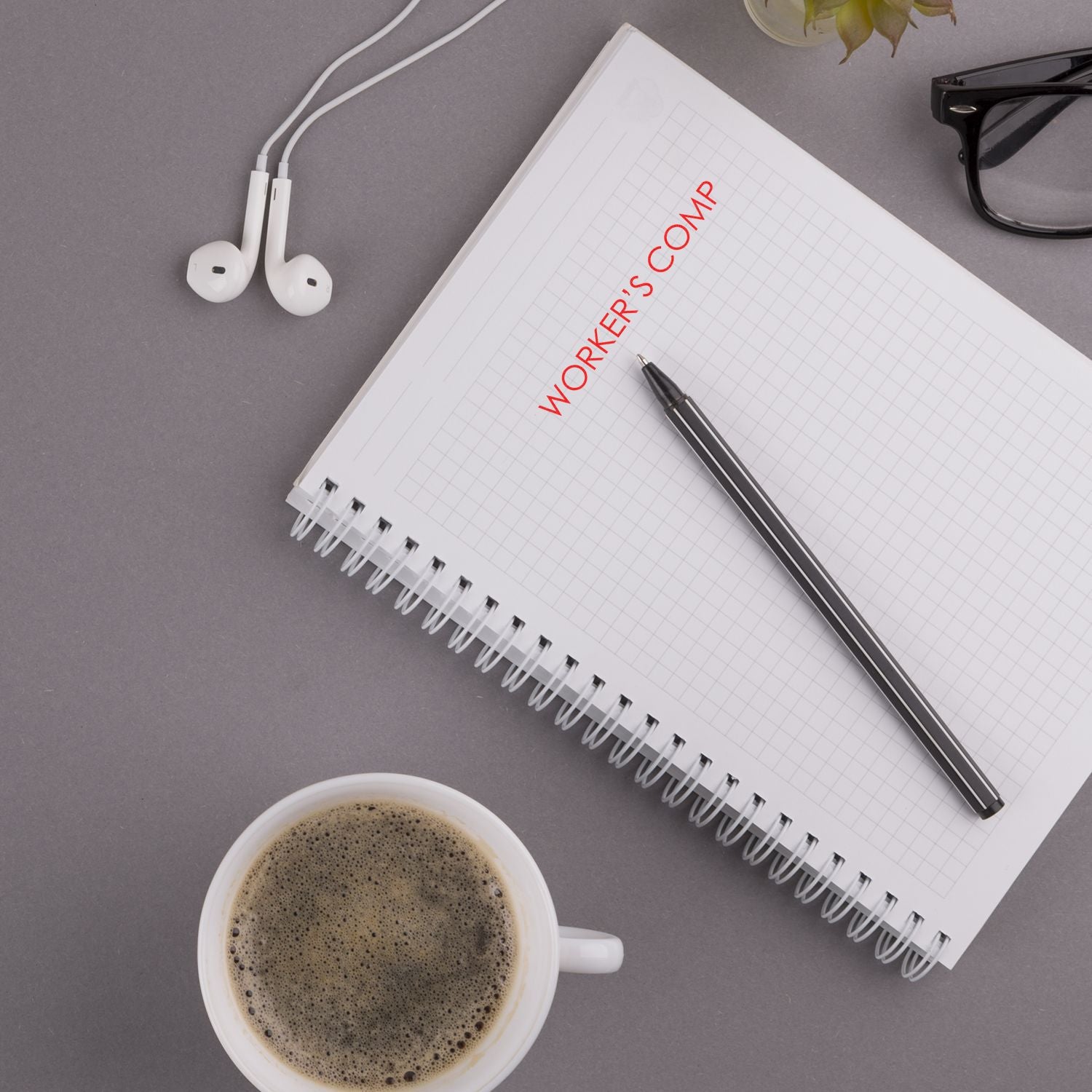 Workers' compensation insurance concept: notebook labeled WORKERS COMP, pen, earbuds, and coffee on an office desk.