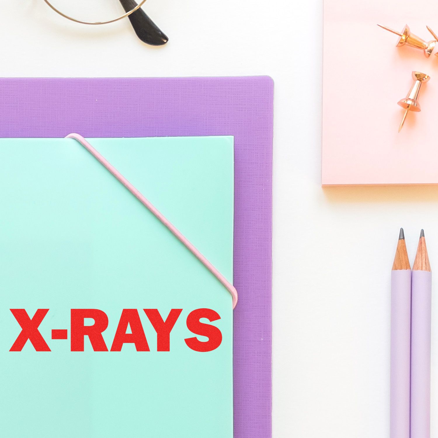 Pastel stationery flat lay with X-rays folder, pencils, push pins, and glasses on a white desk.