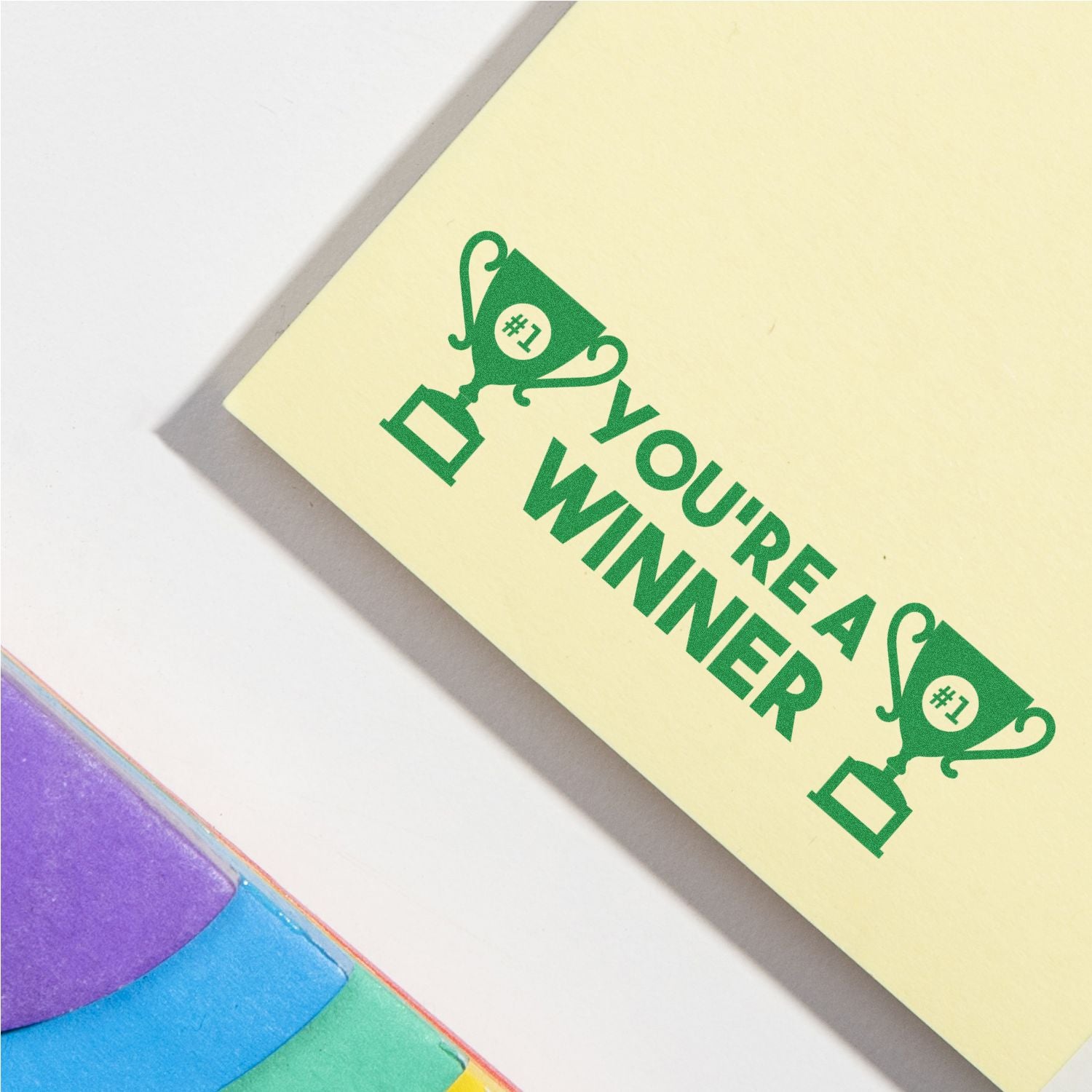 Yellow sticky note with green trophy icon and Youre a Winner text on a pastel background with colorful paper corners.