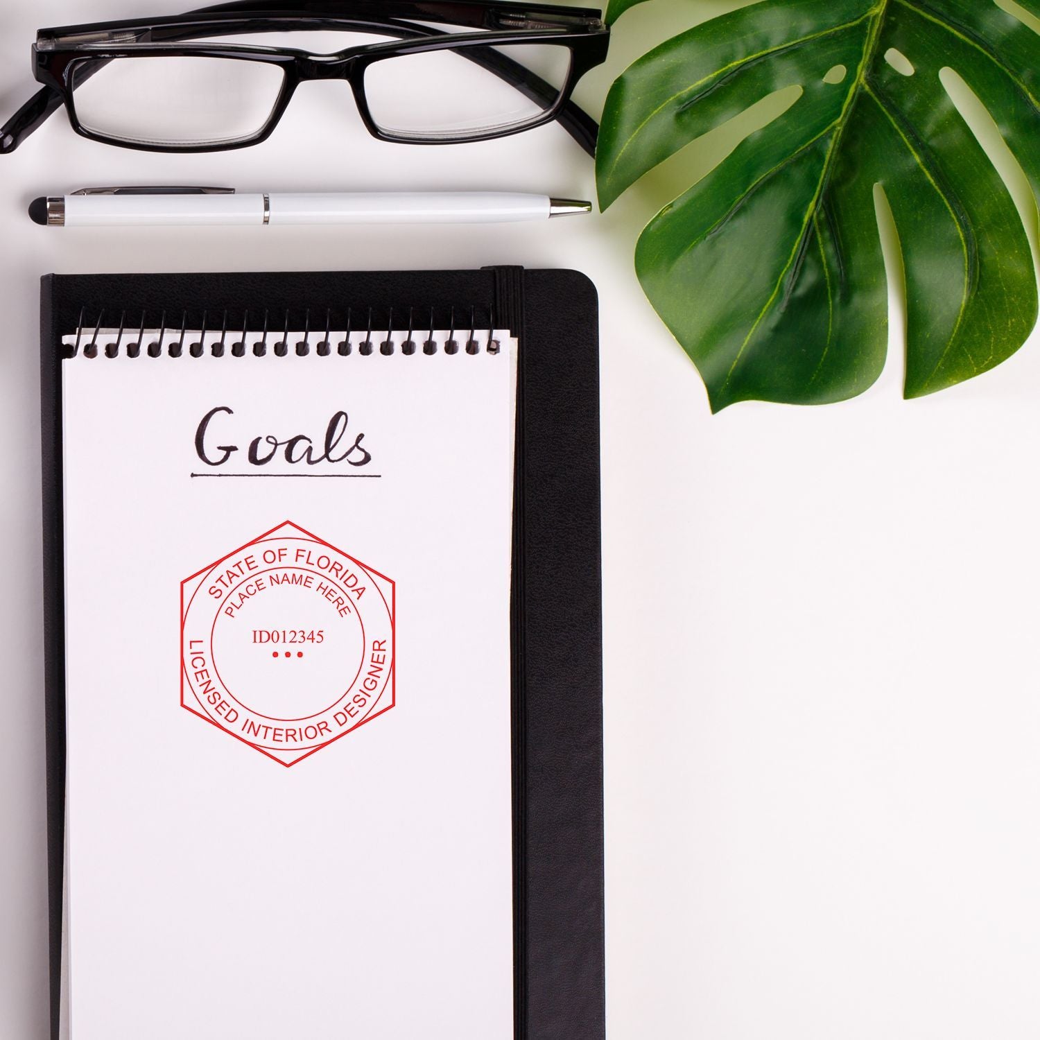 Interior designer goals notebook with glasses, pen, and tropical leaf on a white desk, featuring a Florida licensed interior designer seal.