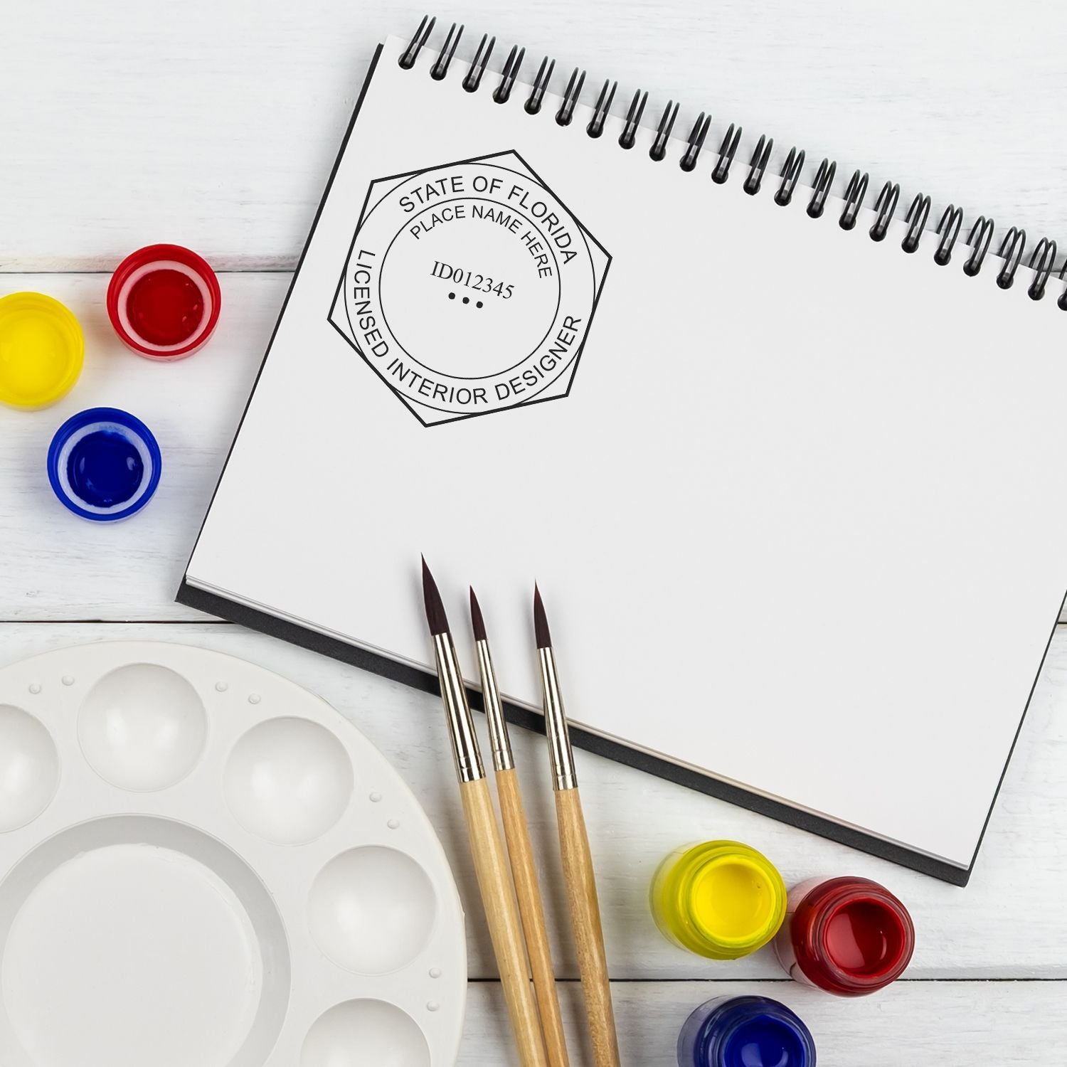 Florida licensed interior designer sketchbook with paint brushes, watercolor palette, and colorful paint jars on a white desk.