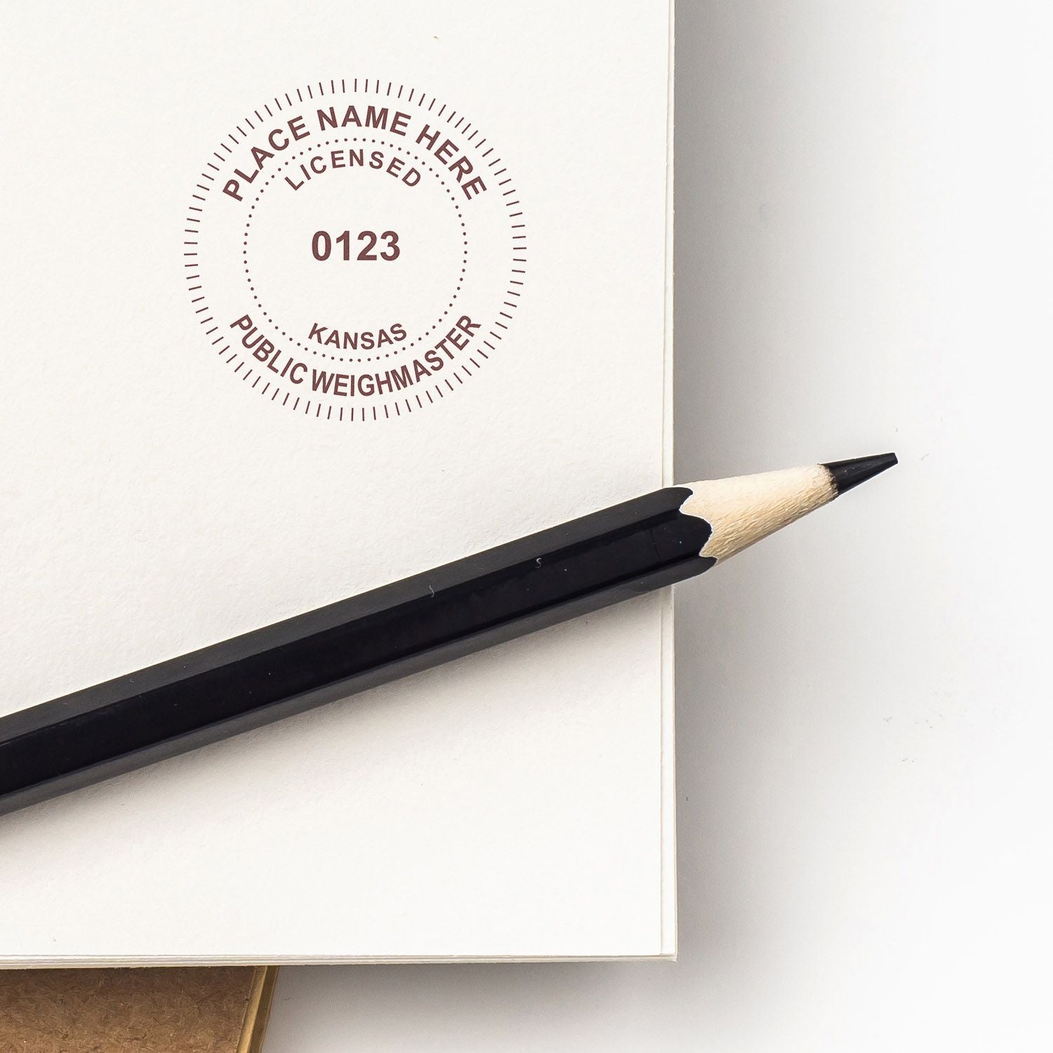 Black pencil resting on open notebook with a circular Kansas public weighmaster stamp design on white paper.
