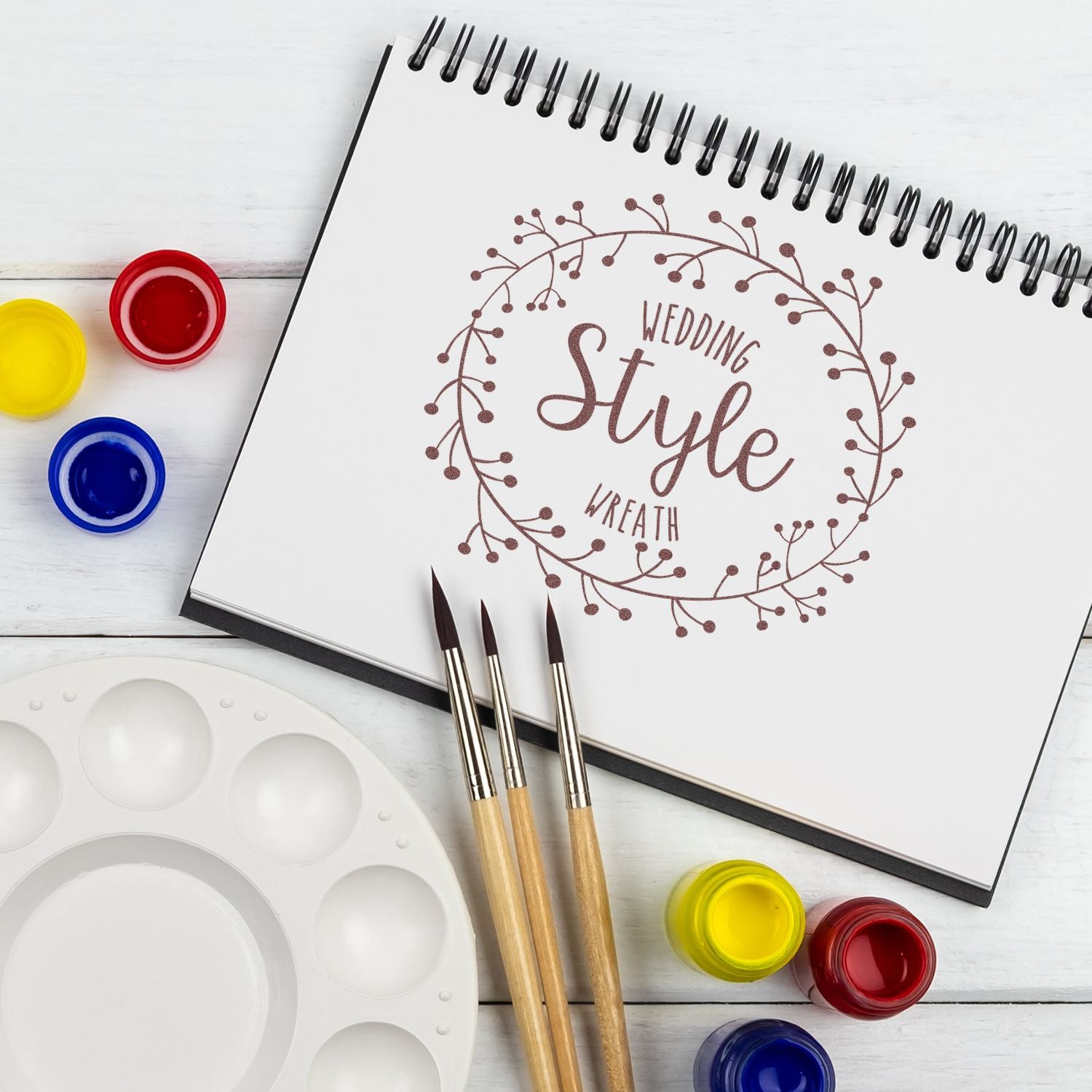 DIY wedding stationery art: sketchbook page with wedding quote lettering, surrounded by paint brushes and colorful paint pots