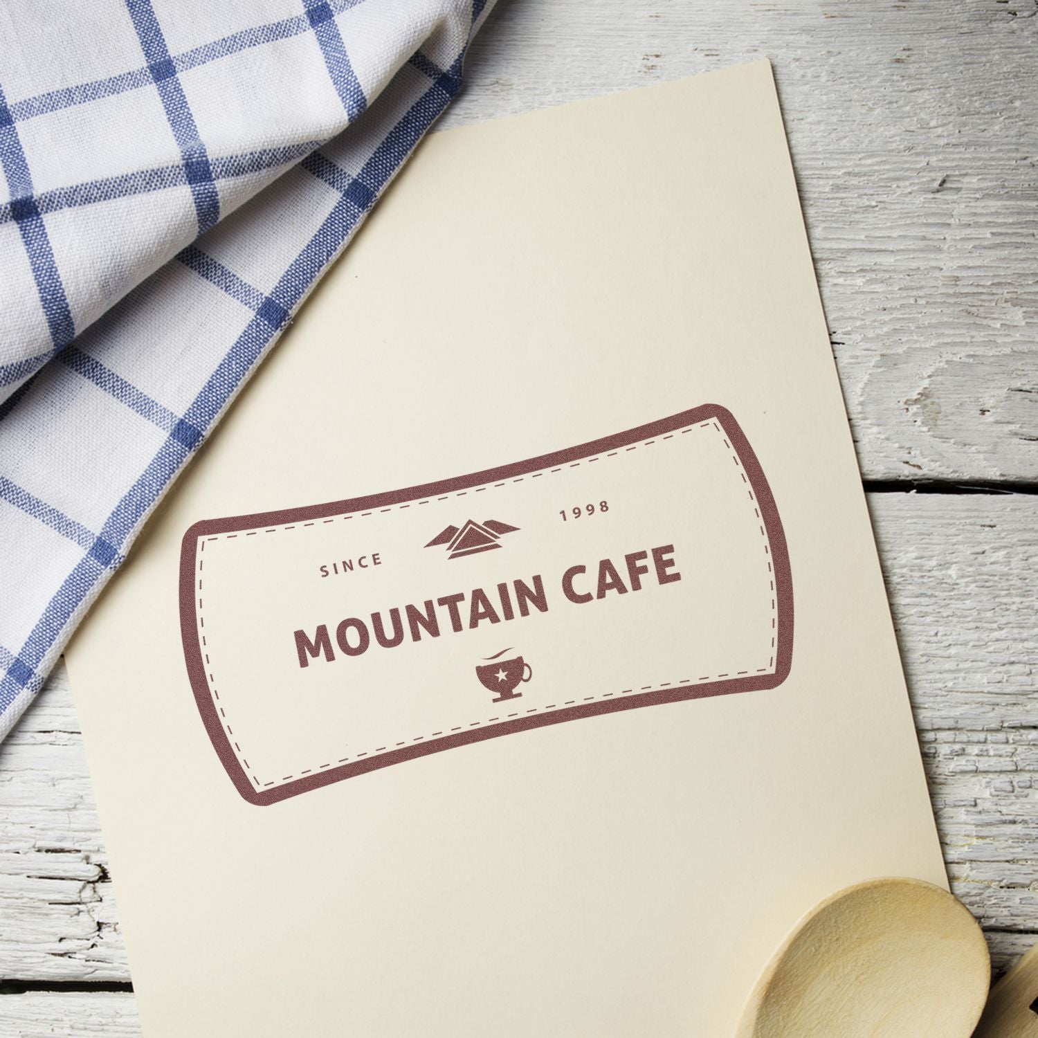 Mountain Cafe branding label on a menu card reading Since 1998, with mountain and trophy icons beside blue checkered cloth.