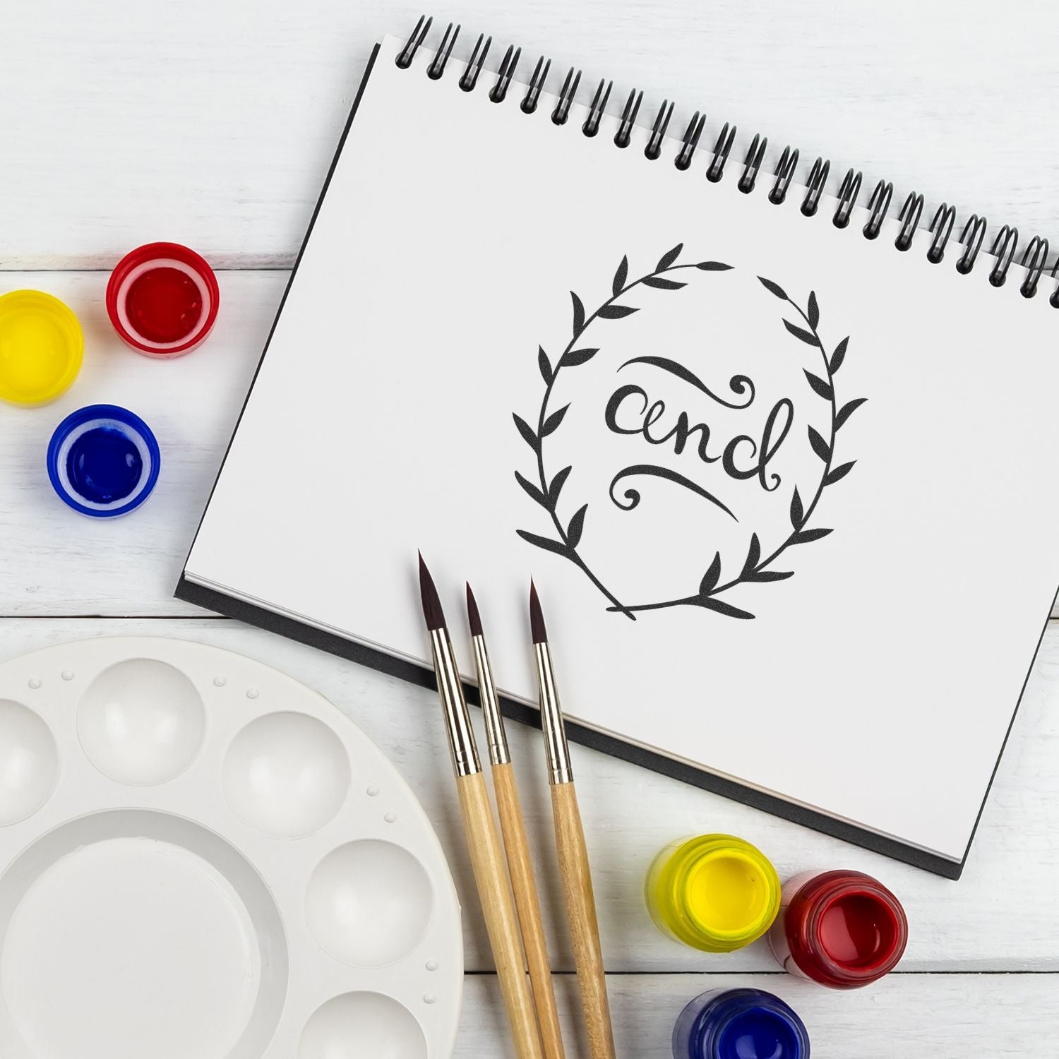 Creative calligraphy art setup: sketchbook page with hand-lettered 'enjoy' laurel, paint pots and brushes on palette, lesson ideas.