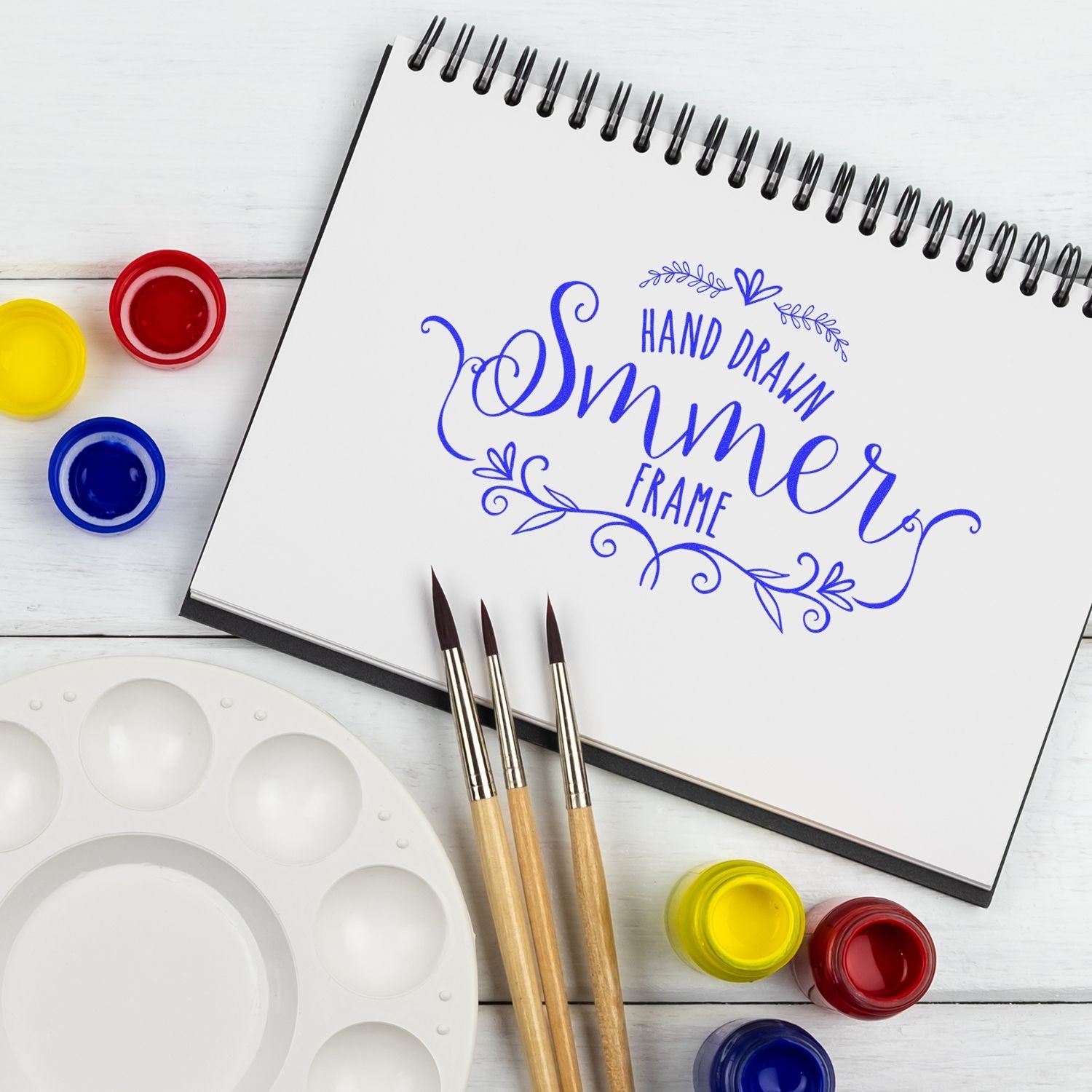Art supplies with open sketchbook showing blue Hand Drawn Frame lettering design and paintbrushes for crafting logo mockup banner.