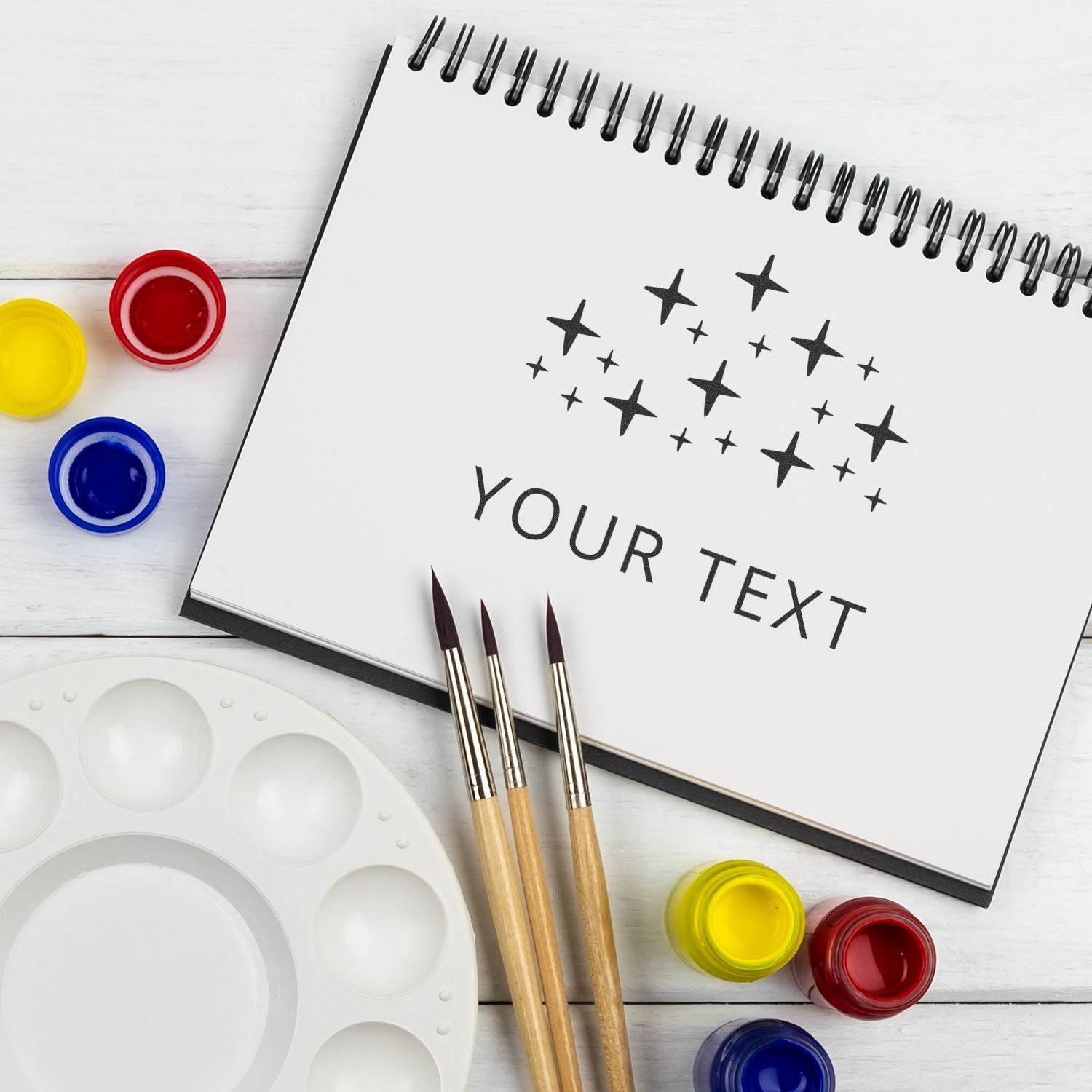 Blank spiral sketchbook mockup with paint brushes, watercolor palette, and colorful paint jars on white wooden table.