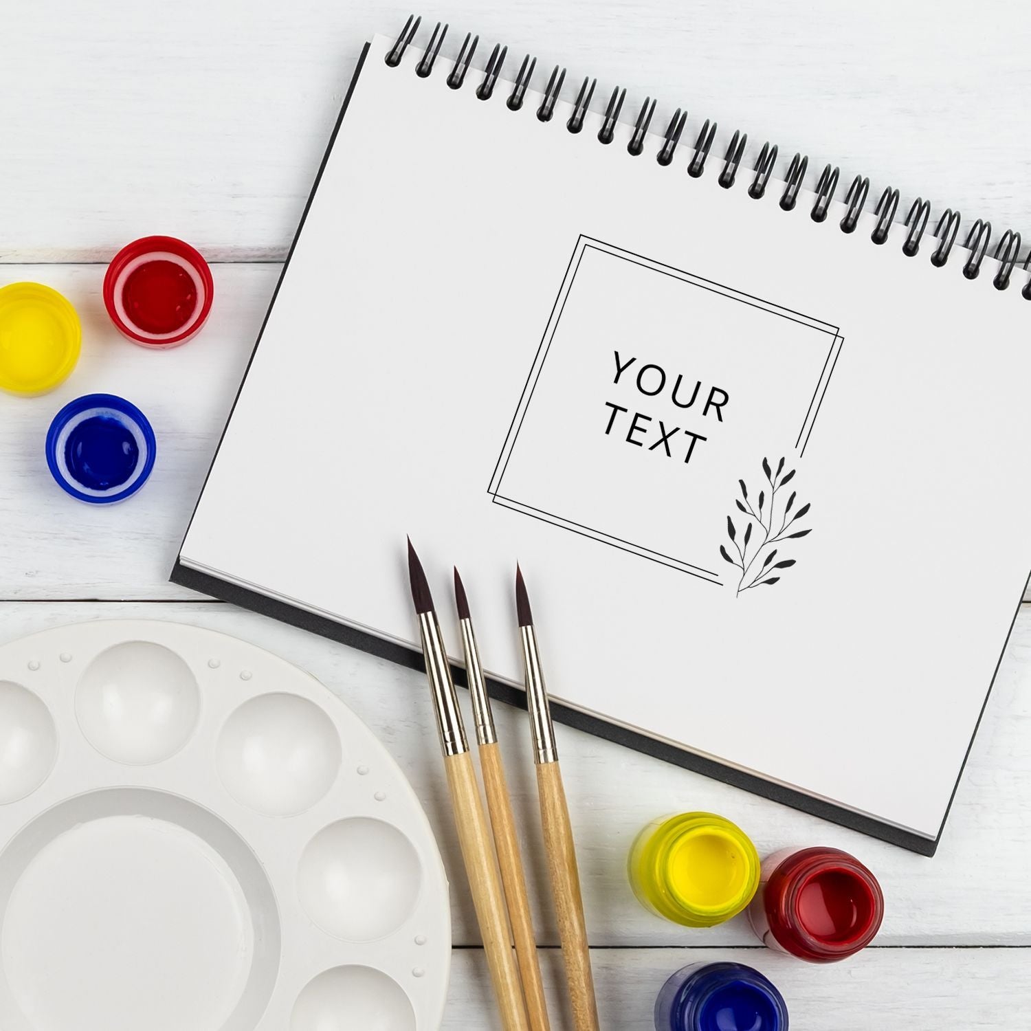Blank sketchbook with YOUR TEXT placeholder, watercolor paints and paintbrushes on a white deskcustom art design space.