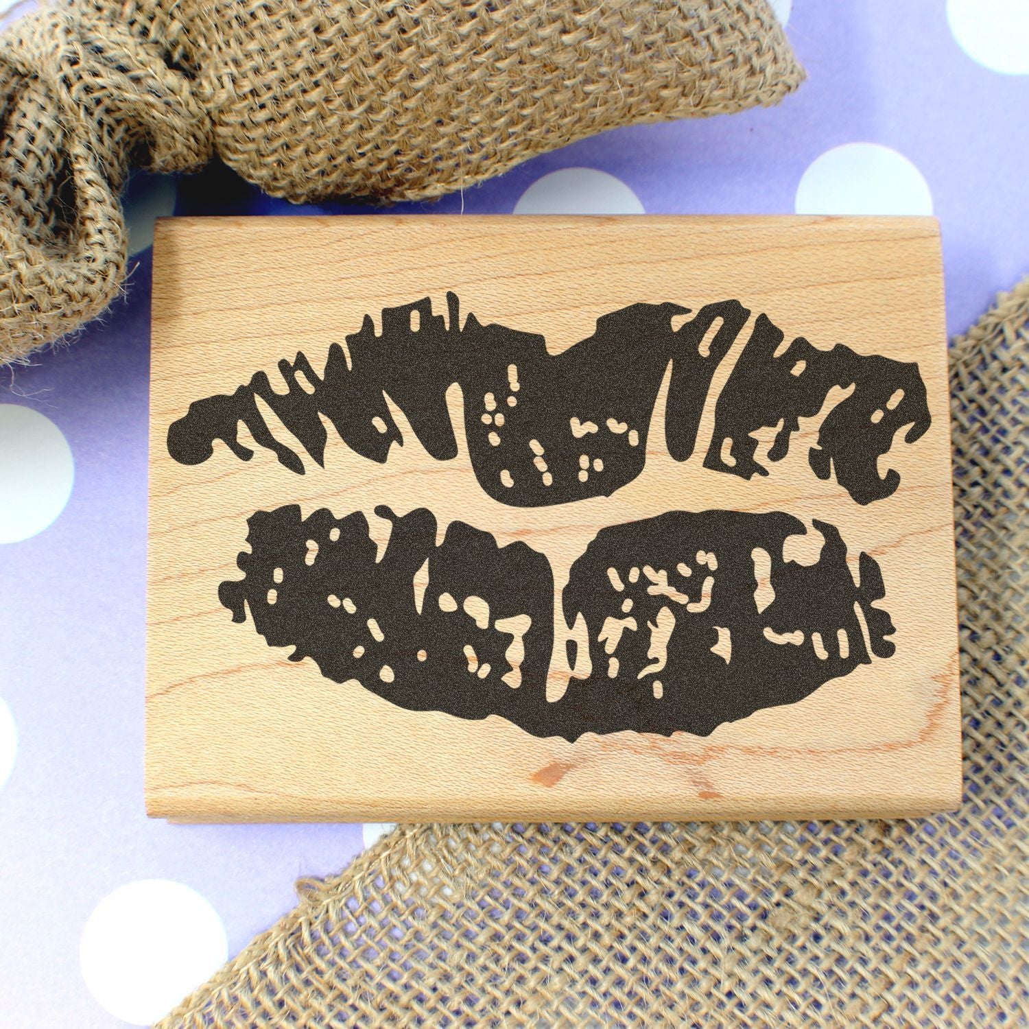 Rustic wooden stamp with black lips silhouette on a pastel polka dot background and burlap fabric accents.