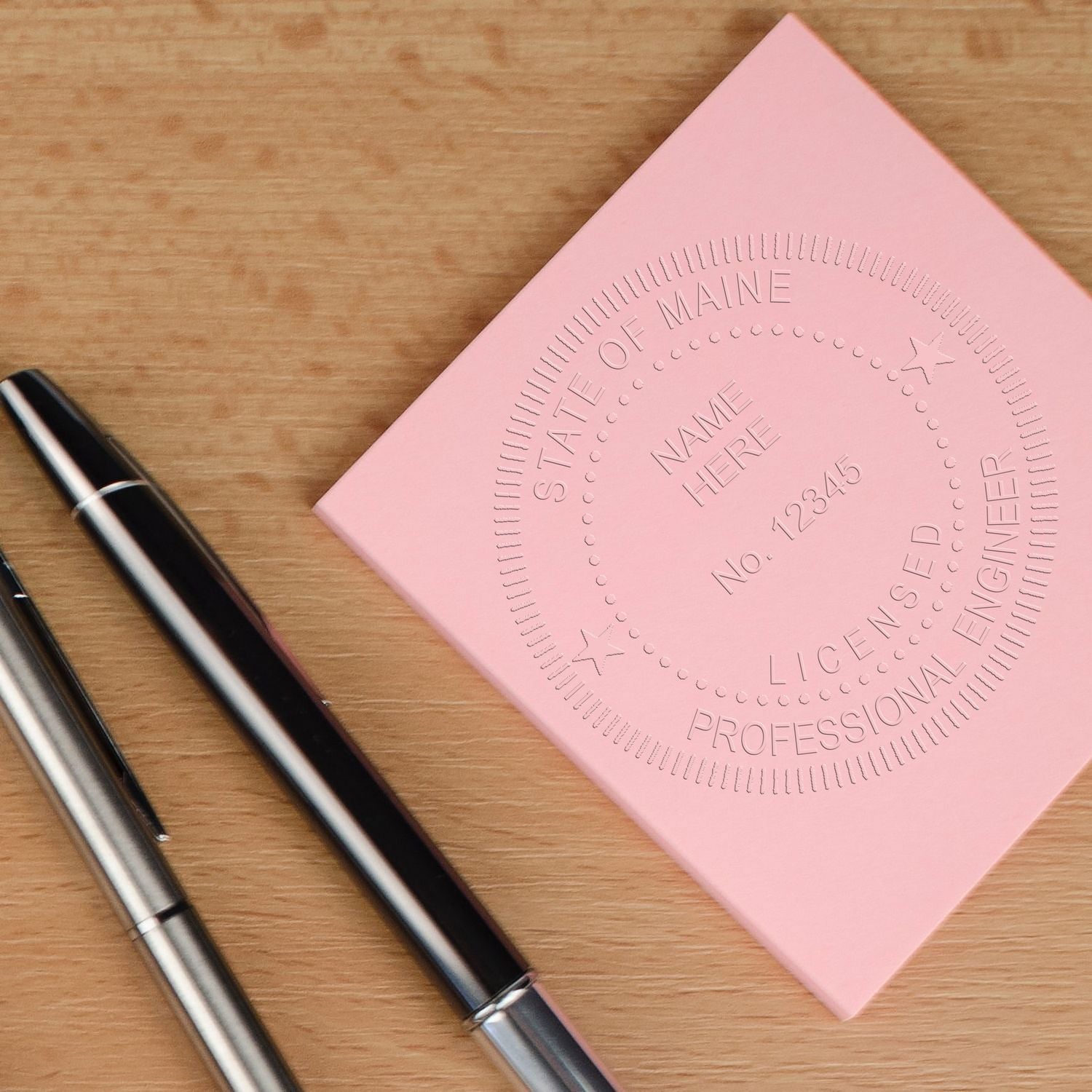 Pink State of Maine professional engineer license stamp card with pens on a wooden desk.