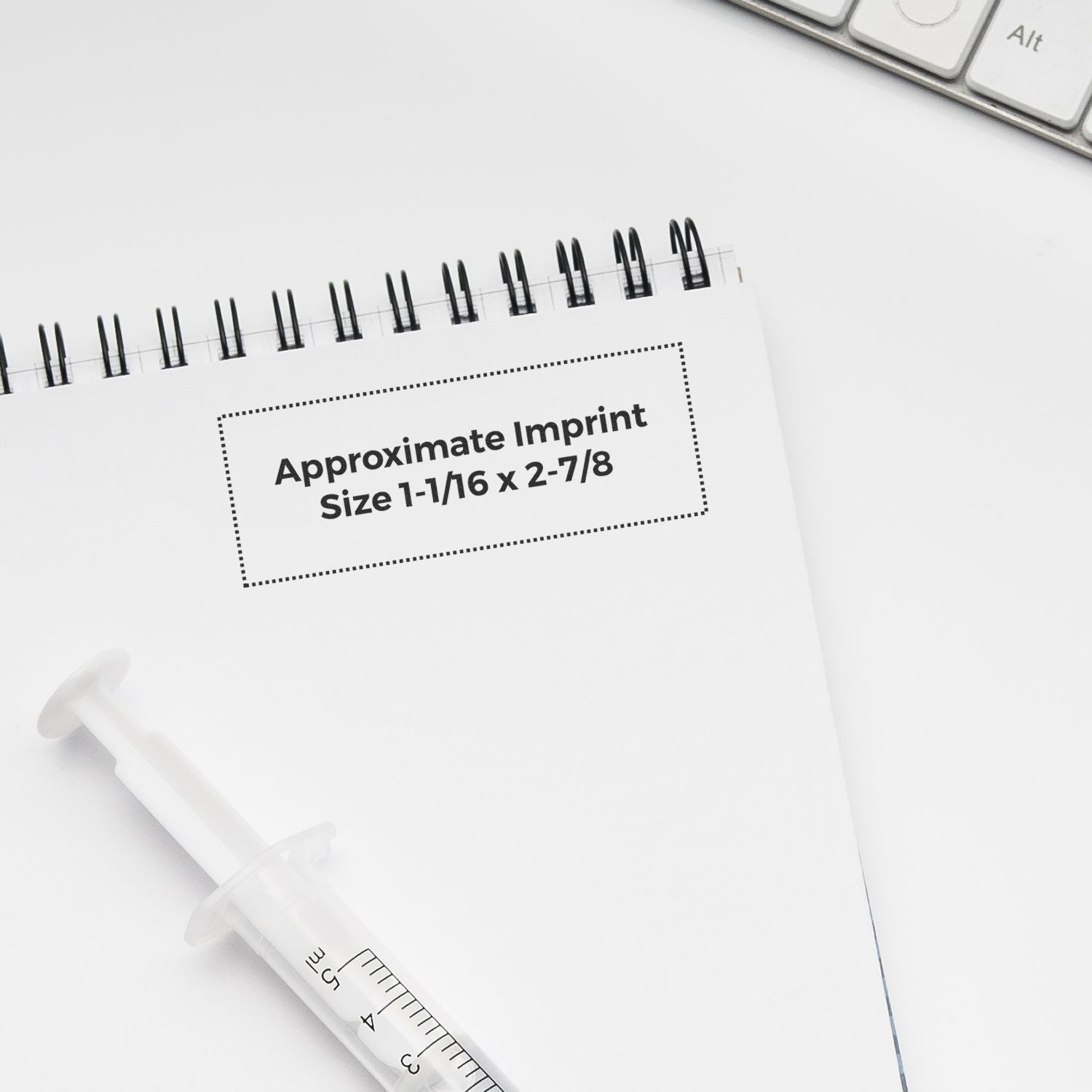 Notebook and syringe on a white desk with Approximate Imprint Size 1-1/16 x 2-7/8 text.