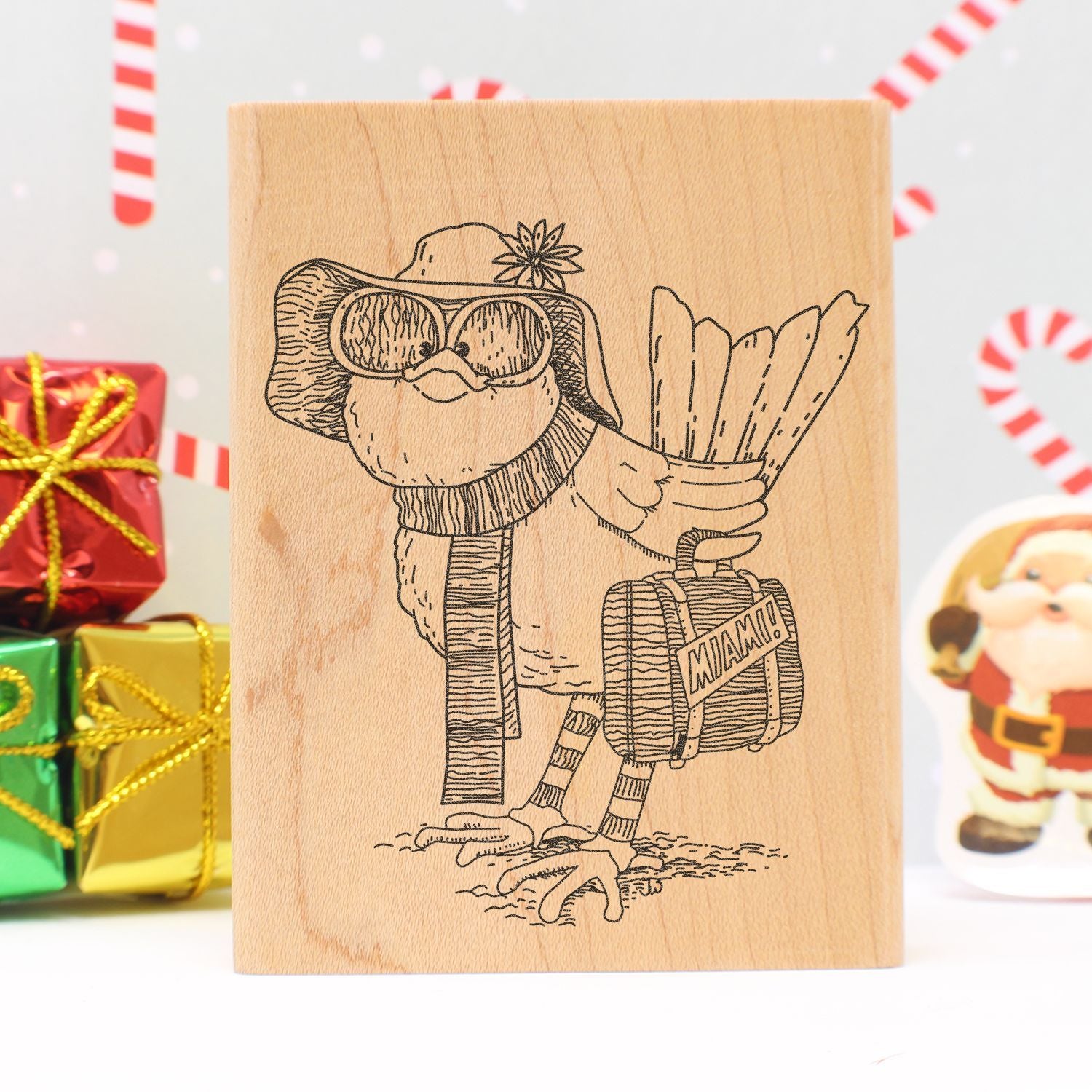 Christmas-themed wooden block print of a whimsical bird wearing winter accessories, holding gifts and a suitcase, with festive presents and Santa decor in the background.