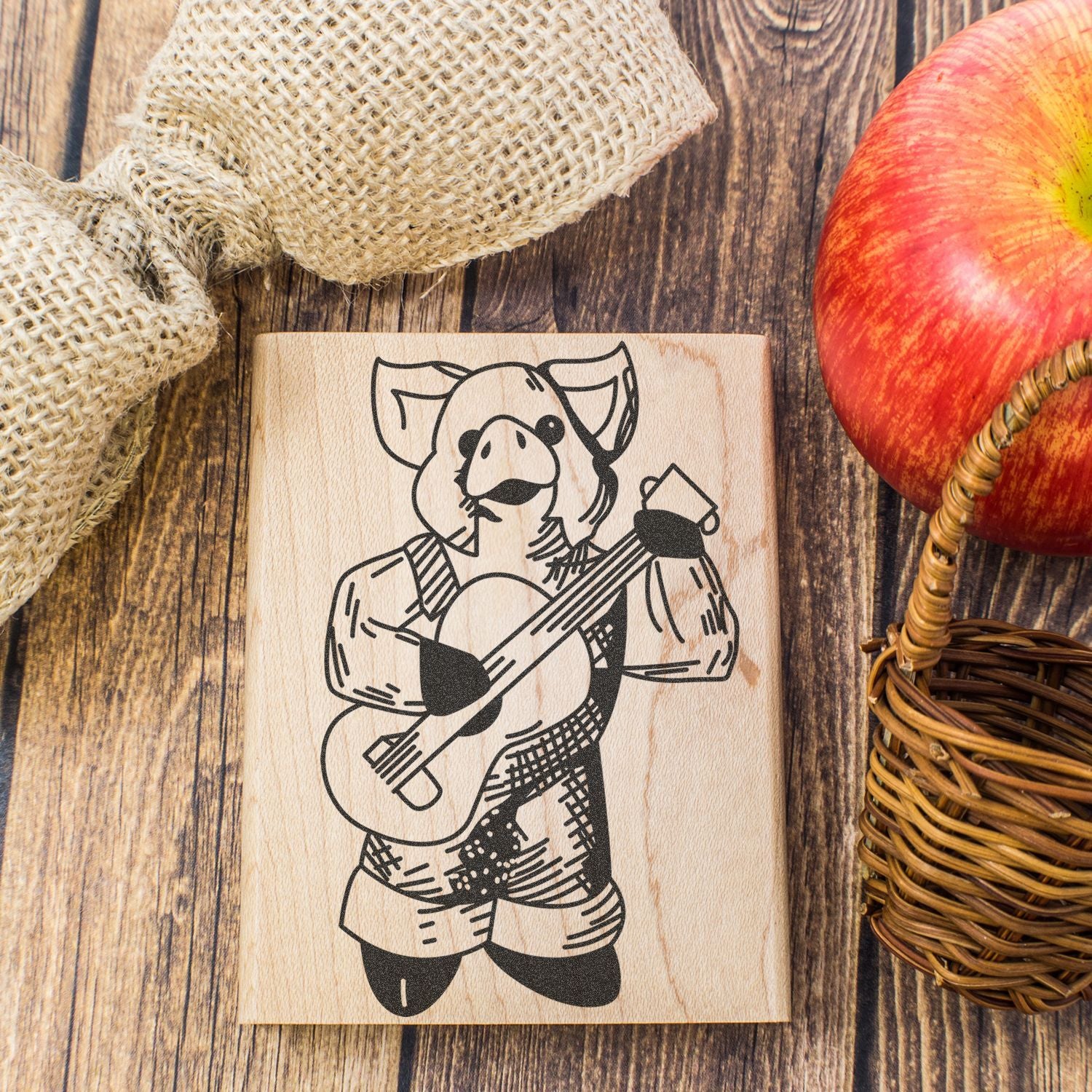 Rustic wooden plaque with a pig playing guitar, styled with burlap and a red apple basket on a wooden table.
