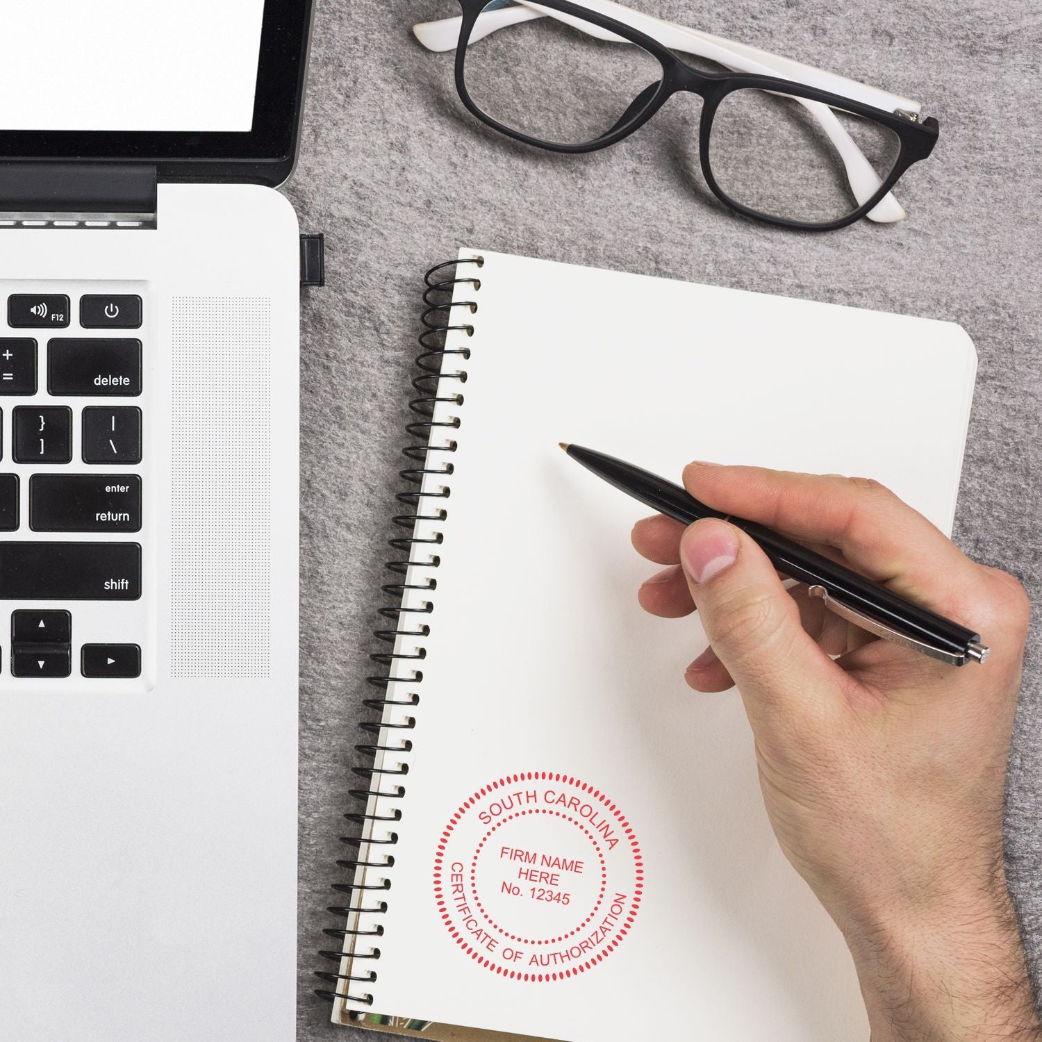 Hand holding pen over blank notebook beside laptop and eyeglasses, with South Carolina certificate of authorization stamp on desk.