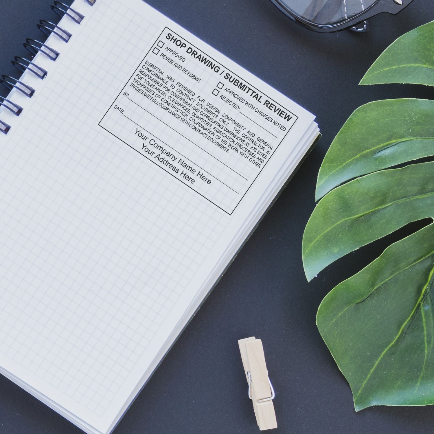 Architectural shop drawing submittal review form in a spiral notebook on a dark desk with green leaves and glasses.