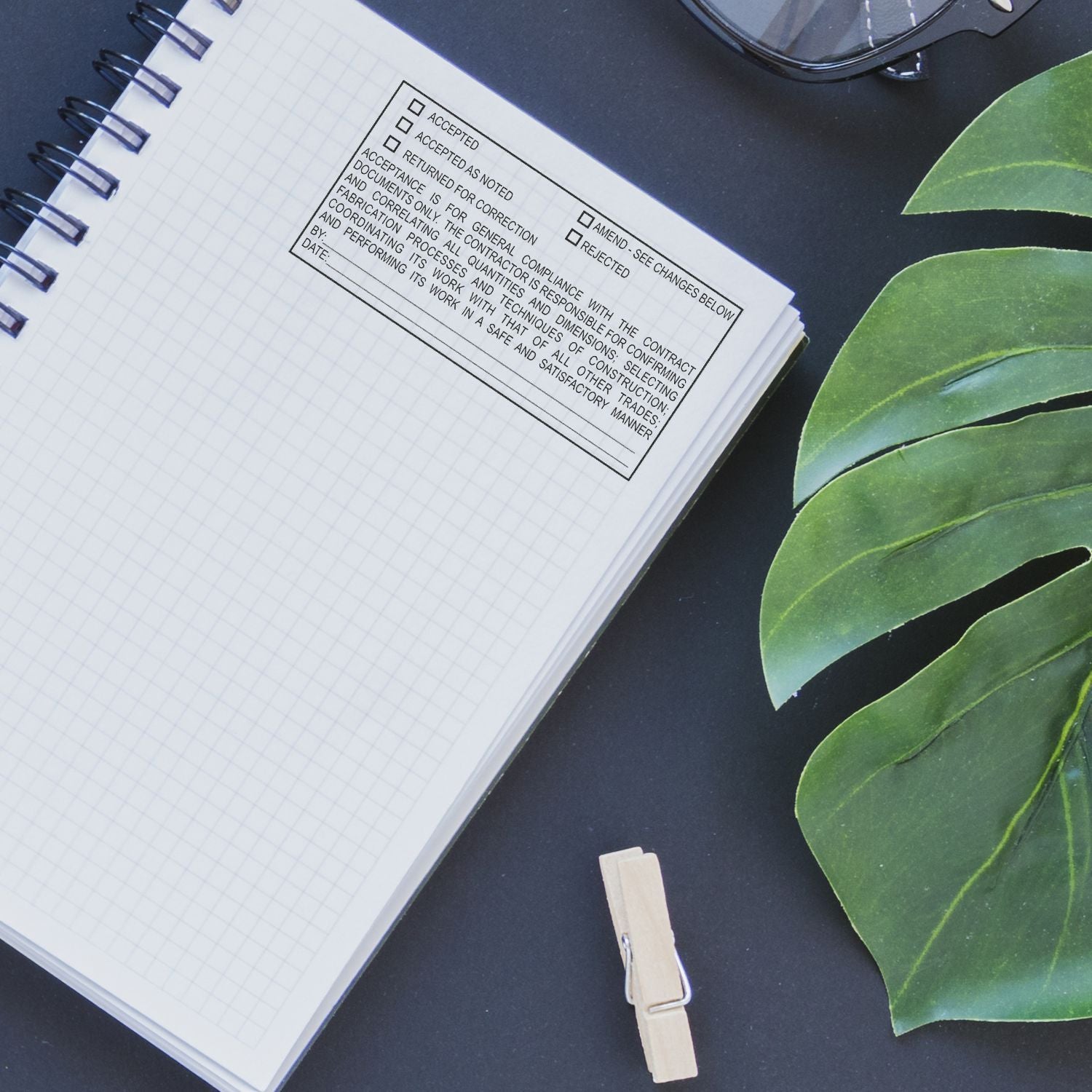 Open notebook with approval stamp, glasses, and green plant leaf on a dark desk, flat lay office workspace concept.