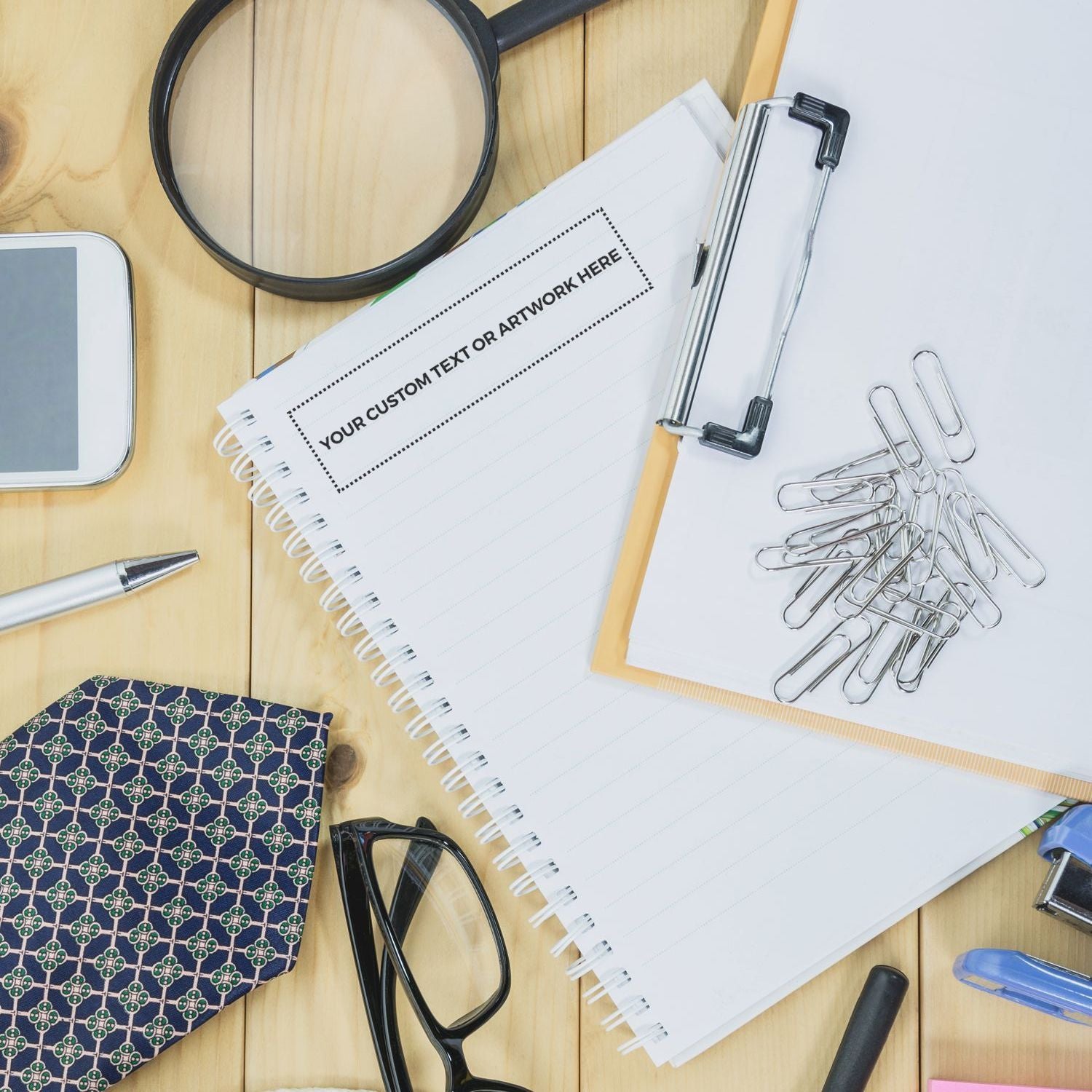 Custom text or artwork placeholder on spiral notebook mockup with stationery tools, magnifying glass, and binder clips.
