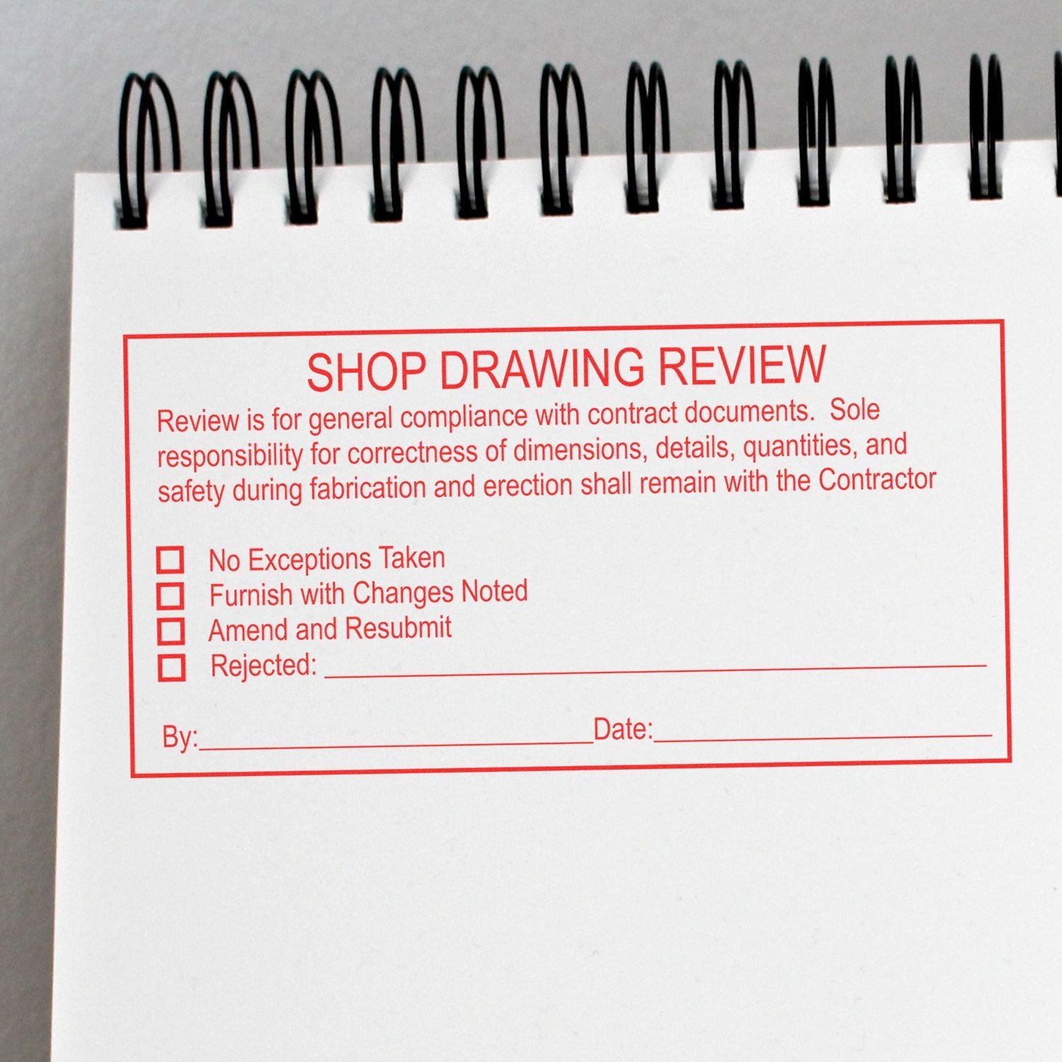 Close-up of a spiral-bound notebook page with a red shop drawing review stamp, including compliance checklist and approval fields.