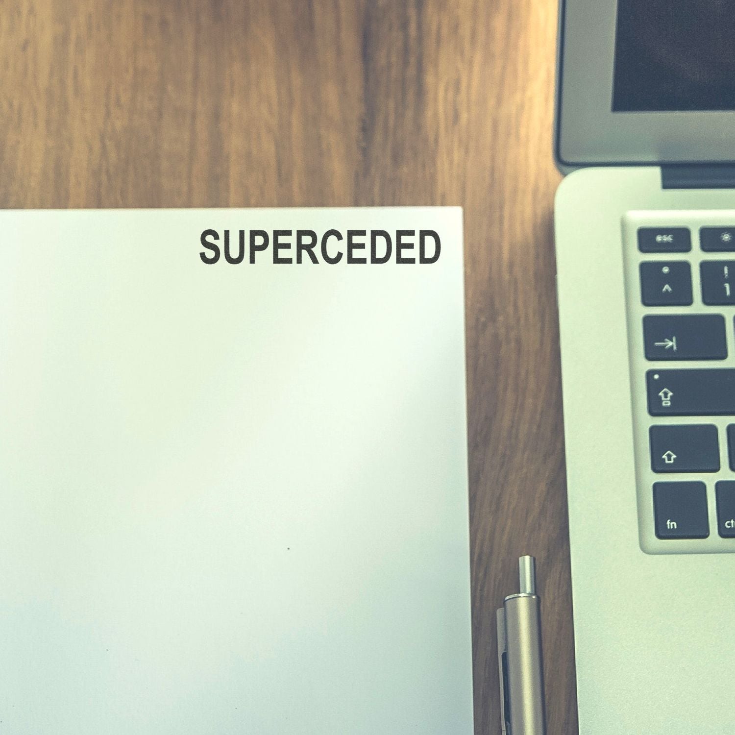 Close-up of a document labeled SUPERCEDED beside a laptop on a wooden desk.