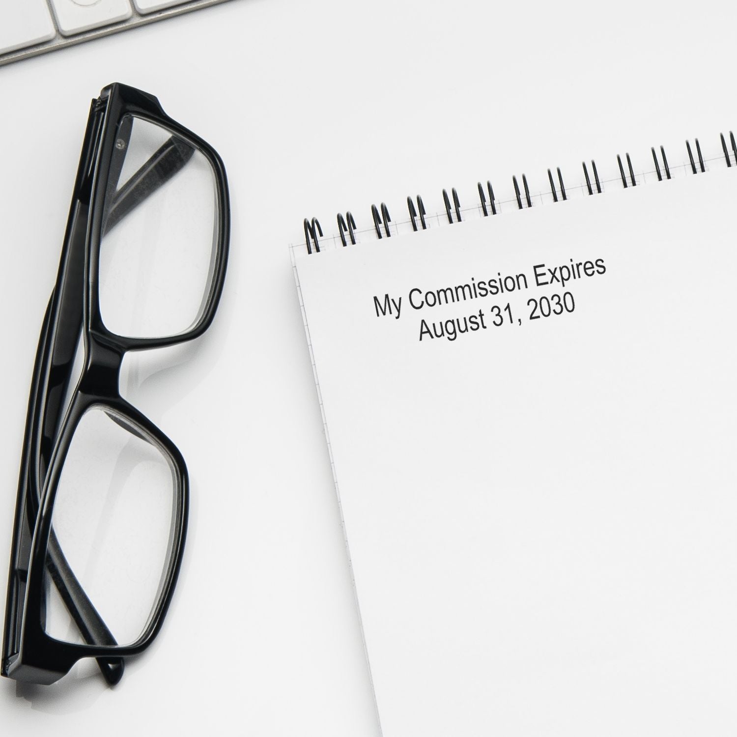 Black eyeglasses beside a spiral notebook with My Commission Expires August 31, 2030 on a clean white desk.