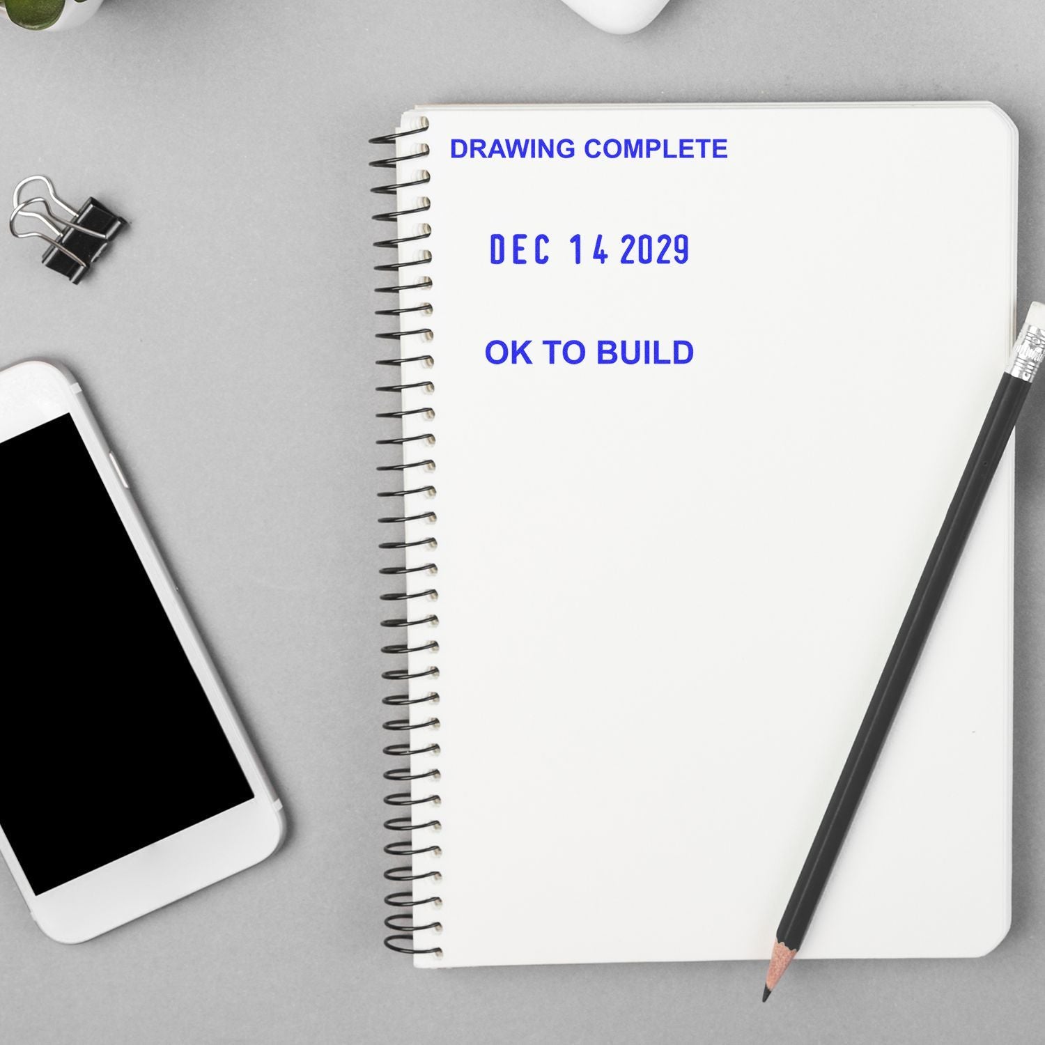 Notebook with Drawing Complete, Dec 14 2029, and OK to Build text beside a pencil, phone, and binder clip on a desk.