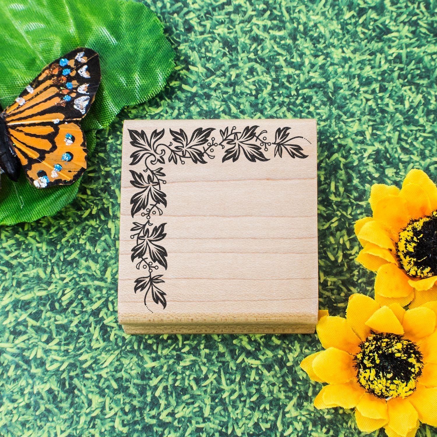 Wooden rubber stamp with leafy floral corner border, butterfly, and sunflowers on a green grass background.