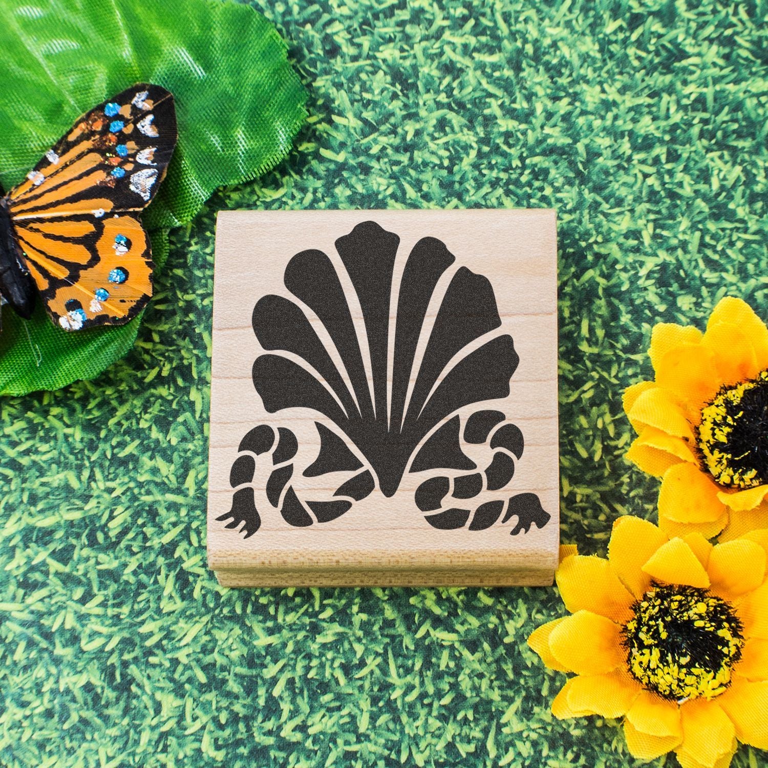 Wooden rubber stamp with floral butterfly design on green grass, surrounded by colorful artificial butterfly and sunflowers
