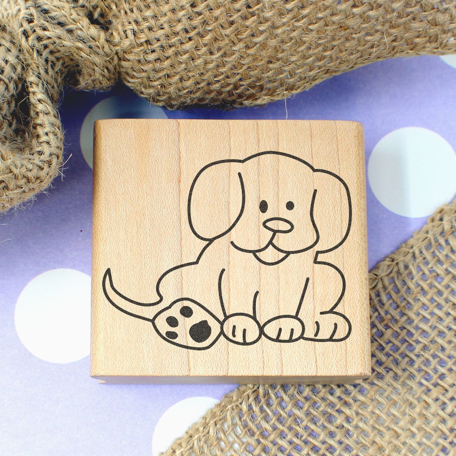 Cute puppy dog wooden stamp with paw print on a rustic wood block, displayed on burlap fabric over a purple polka dot background.