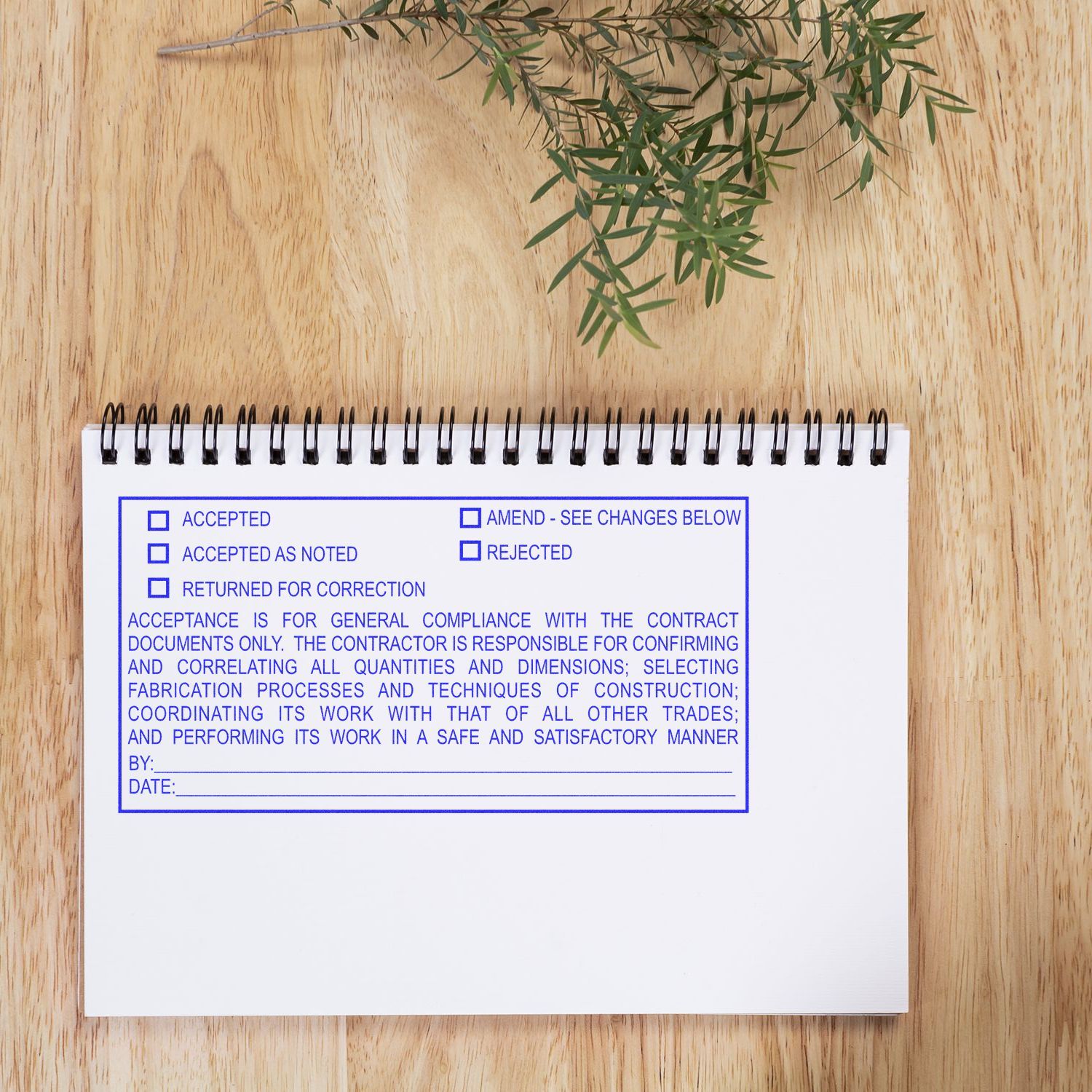 Spiral notebook with contract approval checklist on a wooden desk beside green plant leaves.