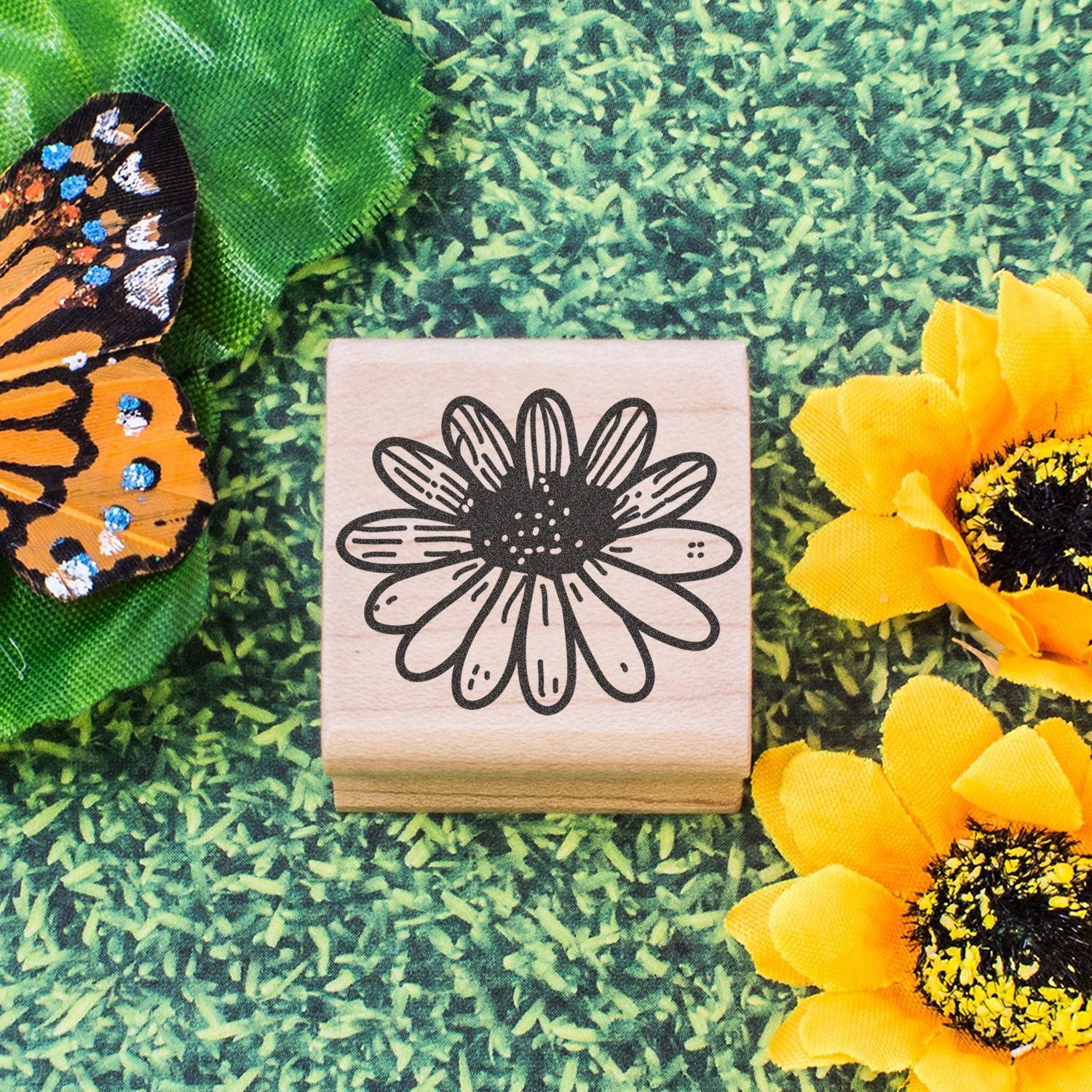Wooden flower stamp with daisy design, butterfly wing, and yellow sunflowers on green grass background.