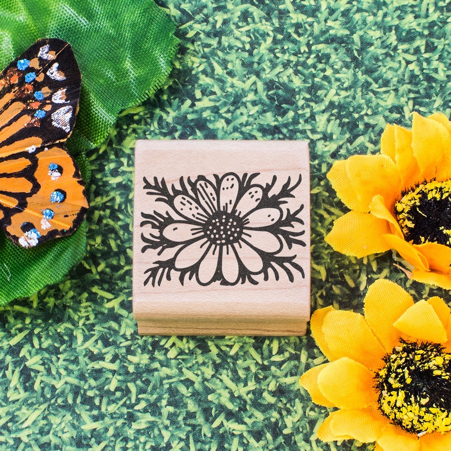 Floral wooden stamp with daisy design, butterfly wing, and yellow sunflowers on green background
