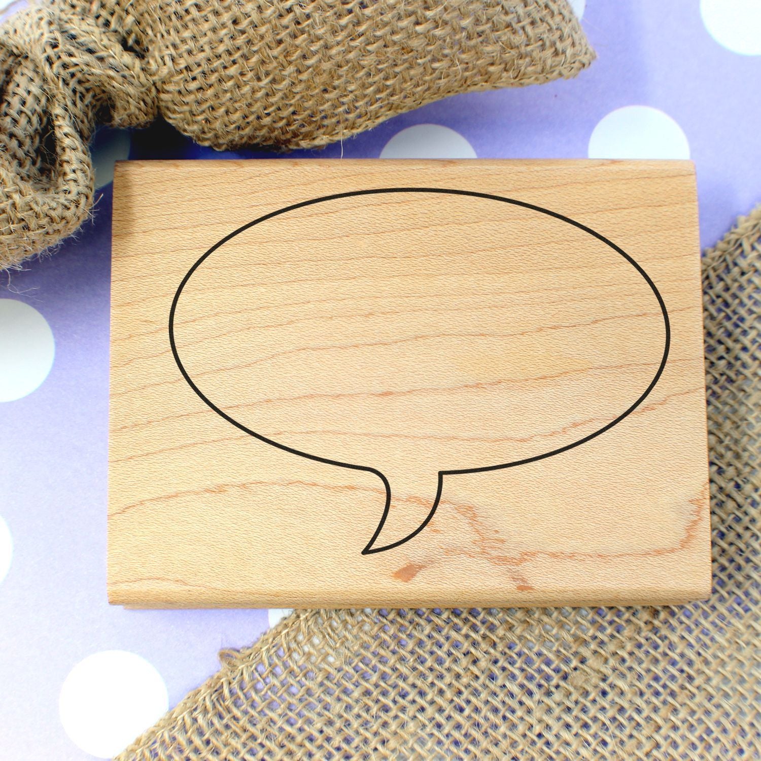 Blank wooden speech bubble sign on burlap fabric with purple polka dot background, rustic decorative message board.