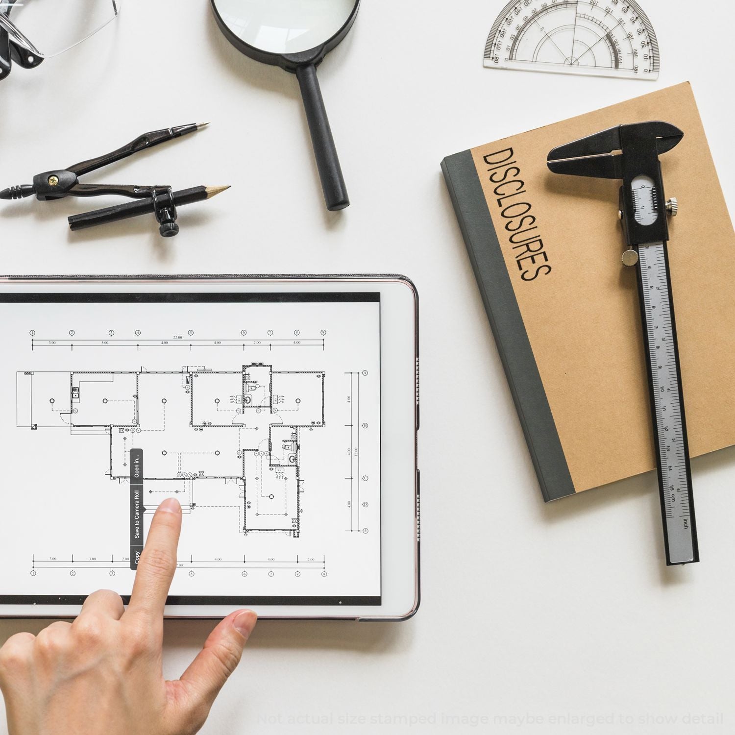 Architectural floor plan on tablet with drafting tools, magnifying glass, compass, and disclosure folder on white desk.