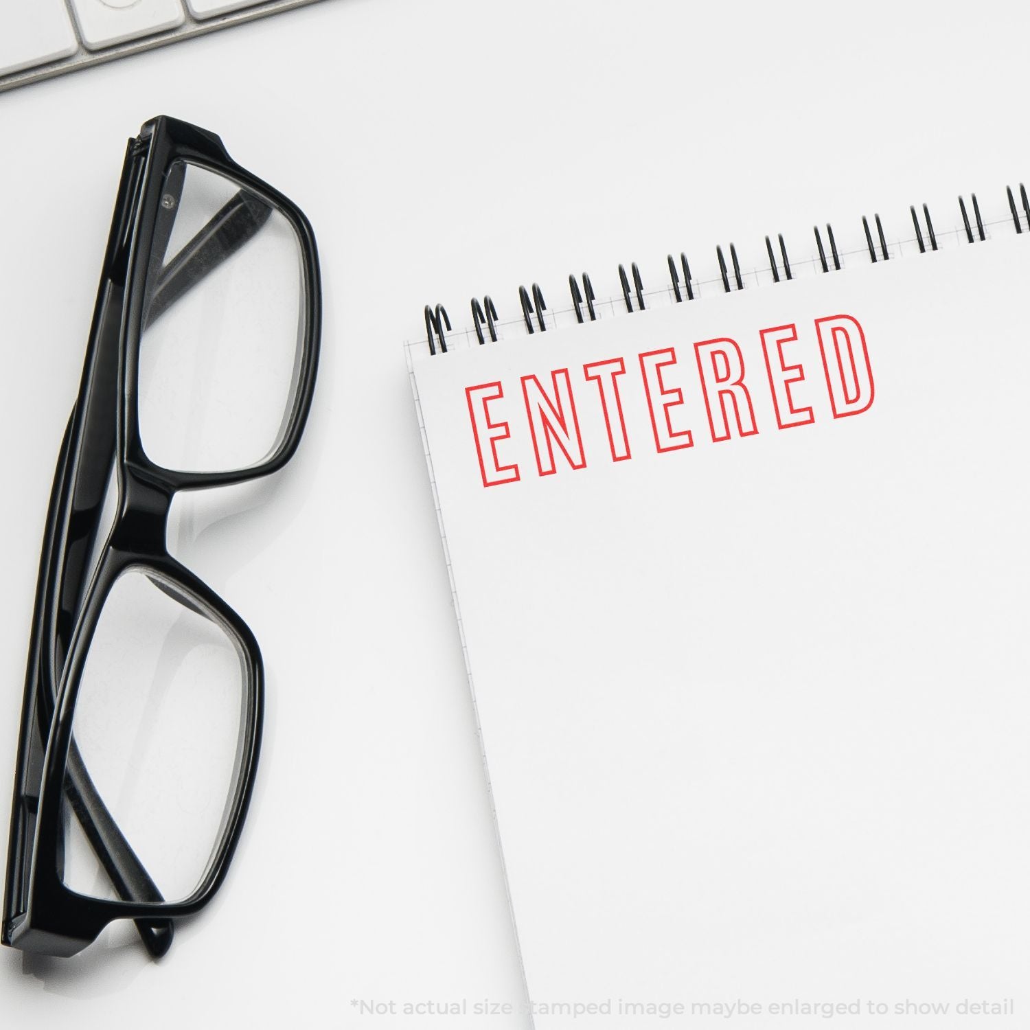 Black eyeglasses beside a spiral notebook stamped ENTERED on a clean white desk.
