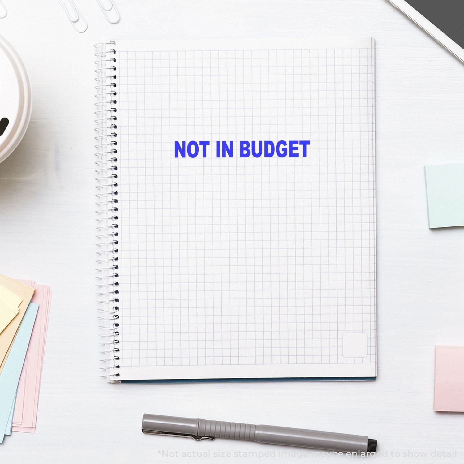 Minimalist notebook with NOT IN BUDGET text on a white desk surrounded by stationery and office supplies.
