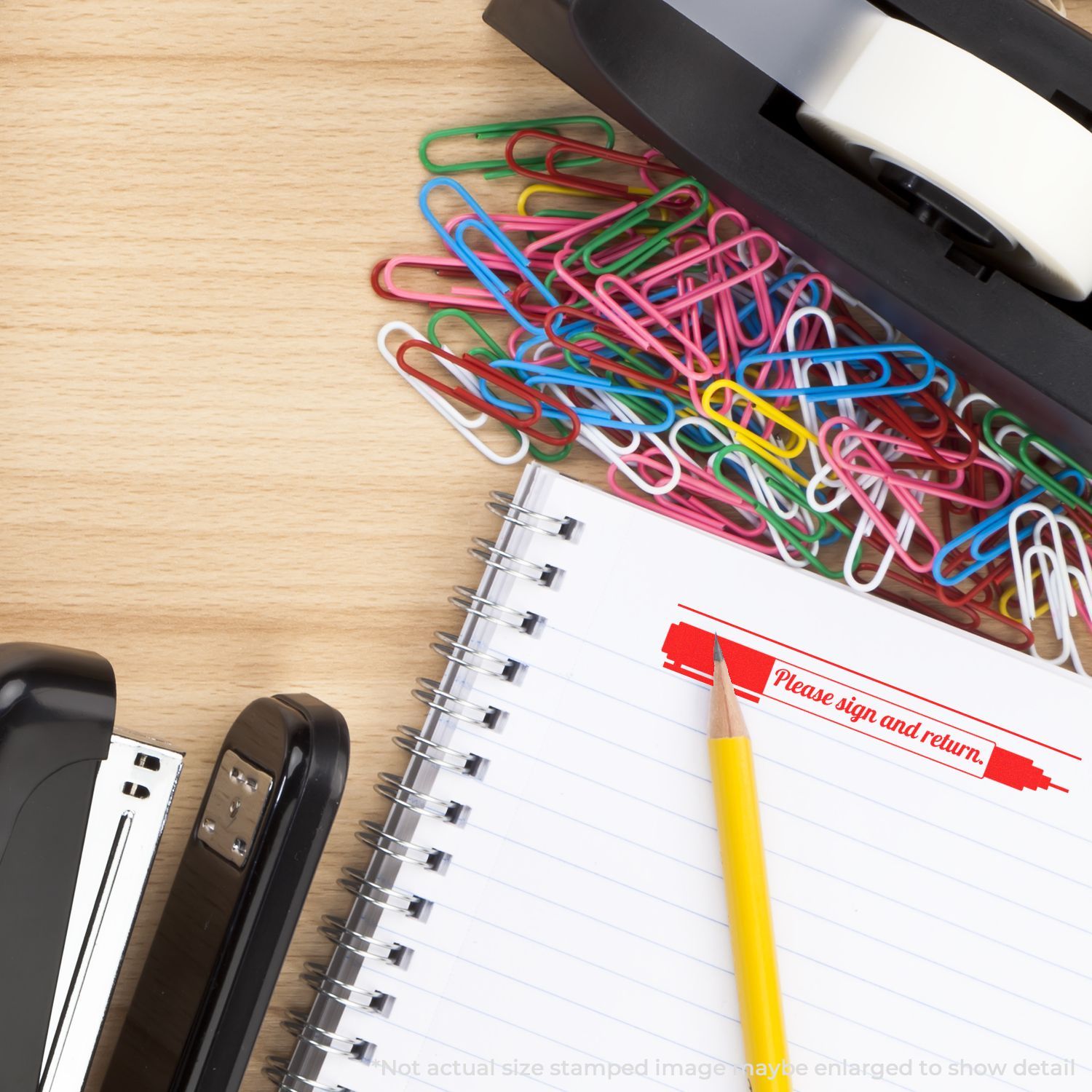 Colorful paper clips, spiral notebook with Please sign and return stamp, pencil, and office supplies on a wooden desk.