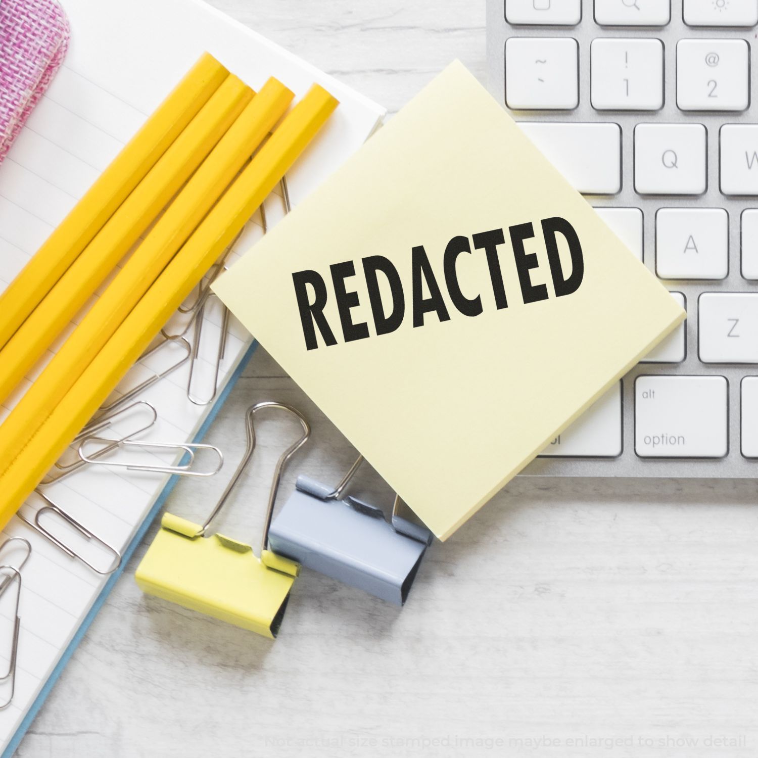 Redacted sticky note on a keyboard with office stationery, yellow pencils, paper clips, and binder clips on a desk.