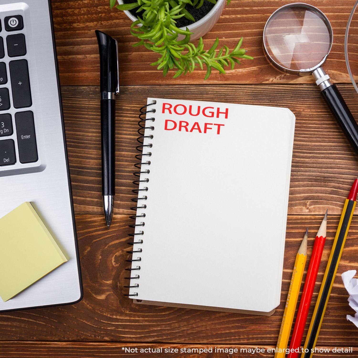 Rough draft notebook on a wooden desk with laptop, pen, magnifying glass, pencils, and plant, representing writing, editing, and creative workspace.