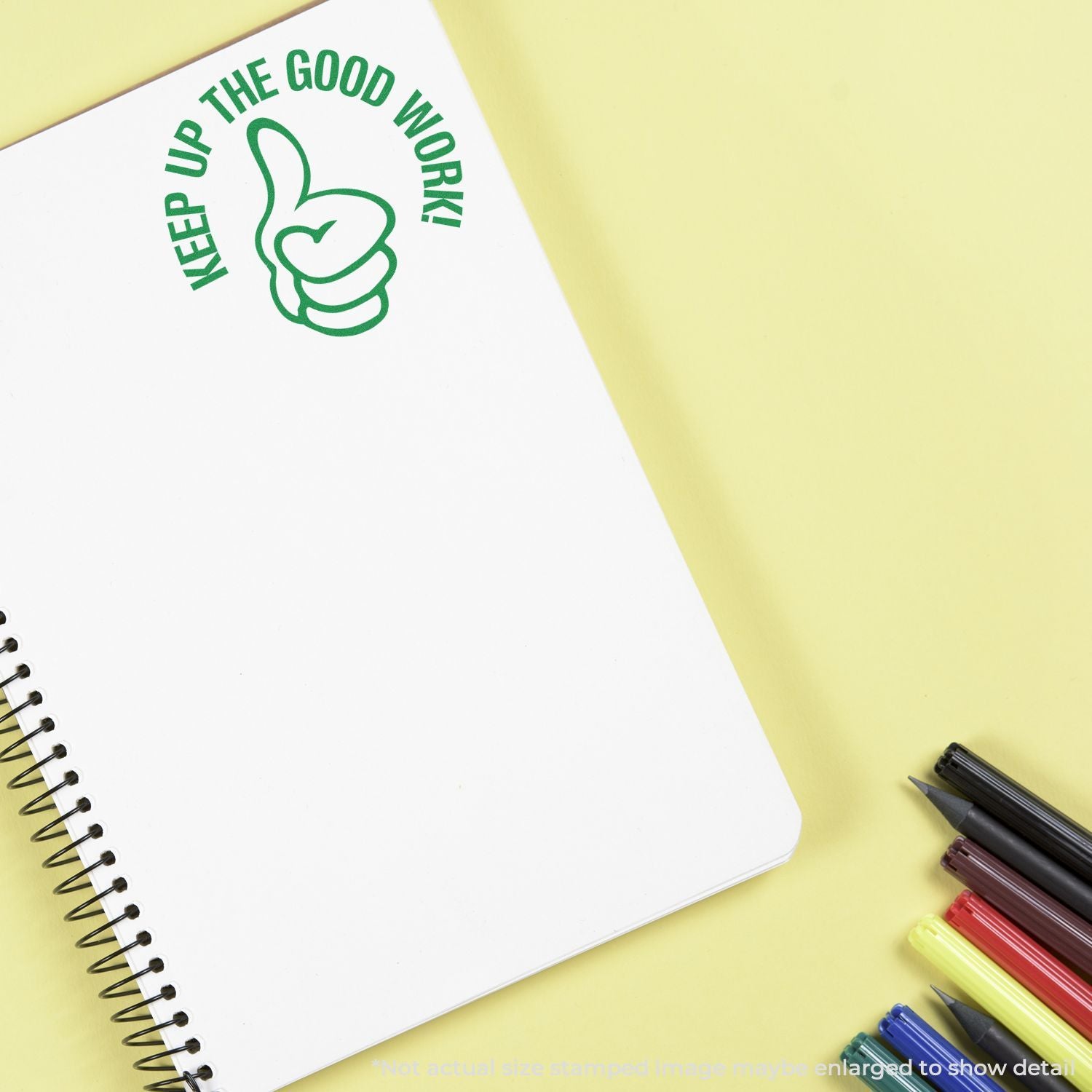 Blank spiral notebook with Keep Up The Good Work thumbs-up graphic, surrounded by colorful pens on a yellow background.