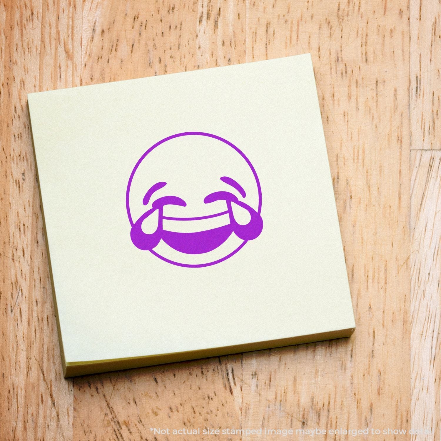 Purple laughing emoji logo printed on a square sticky note on a wooden background.