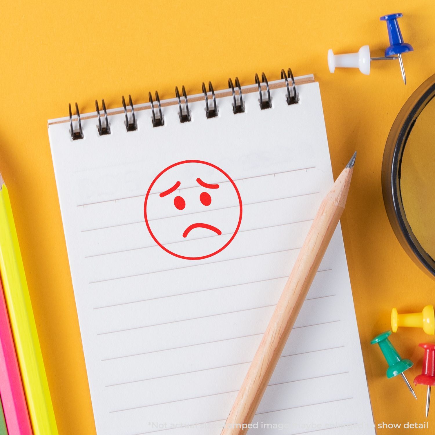 Sad face drawn on a spiral notebook with pencil and colorful push pins on a yellow background, school supplies flat lay.