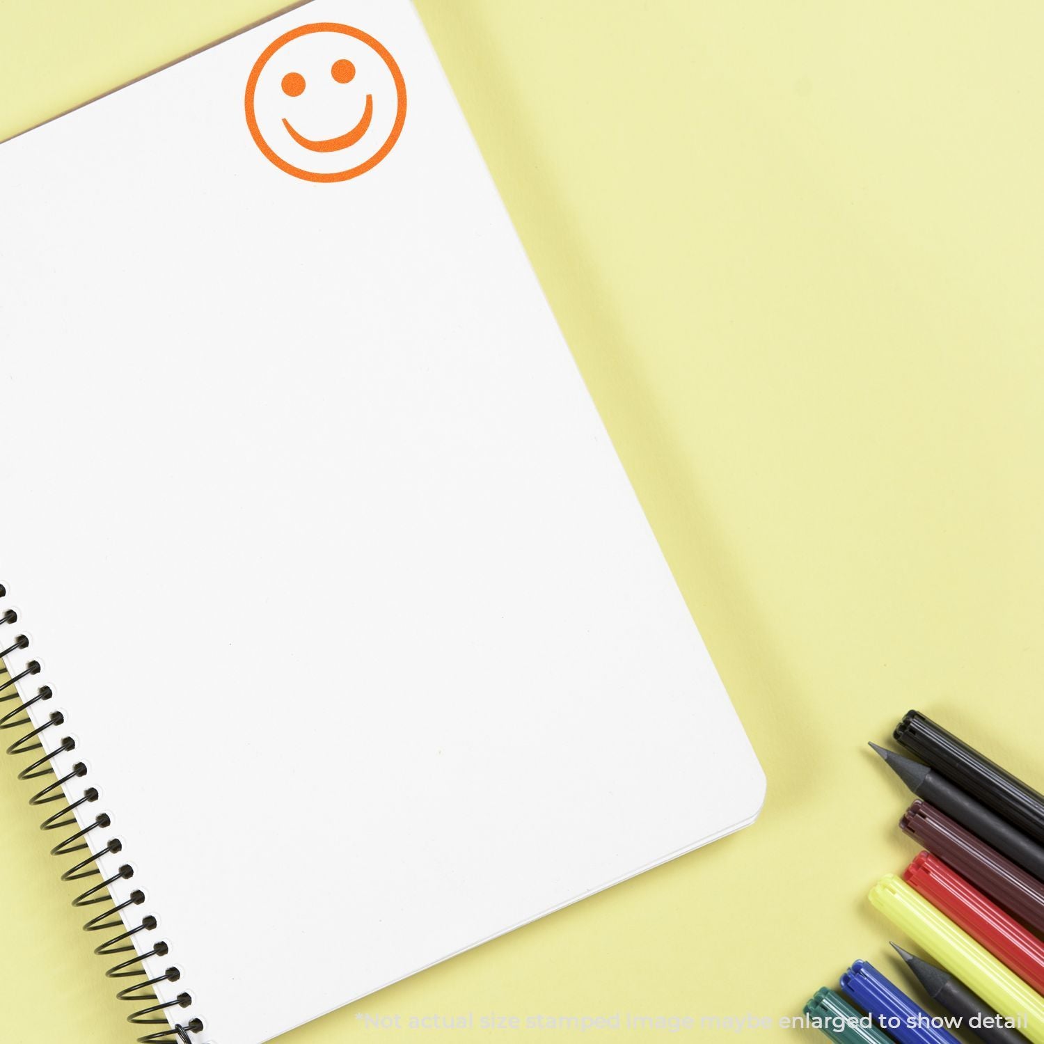 Blank spiral notebook with smiley face stamp on yellow background, surrounded by colorful markers and pens, creative stationery flat lay.