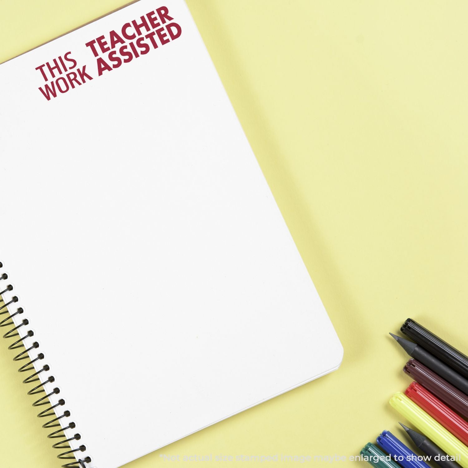 Notebook with This Teacher Work Assisted stamp on yellow background, surrounded by colorful pens and markers.