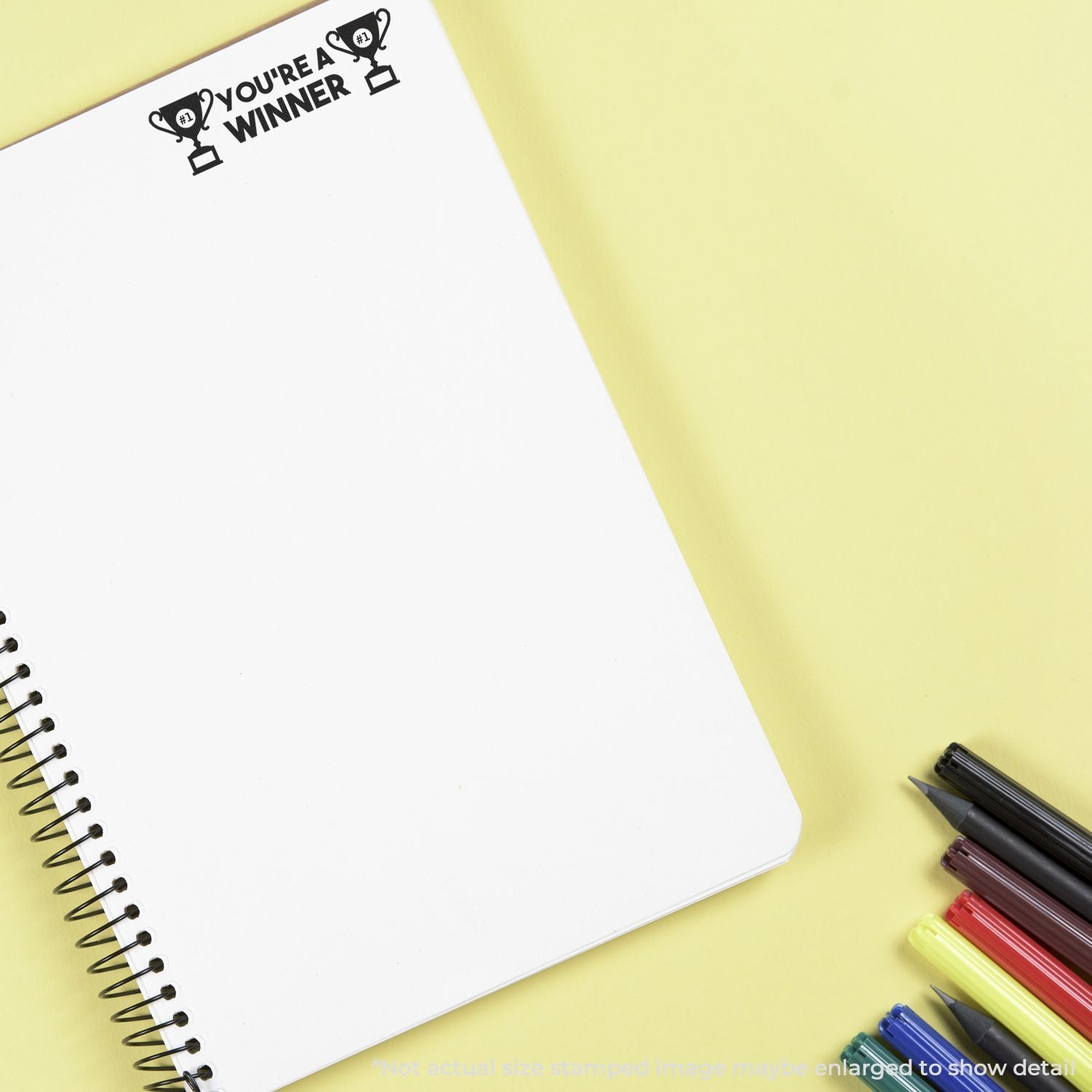 Blank spiral notebook with Youre a winner trophy stamp and colorful markers on yellow background, creative stationery flat lay.