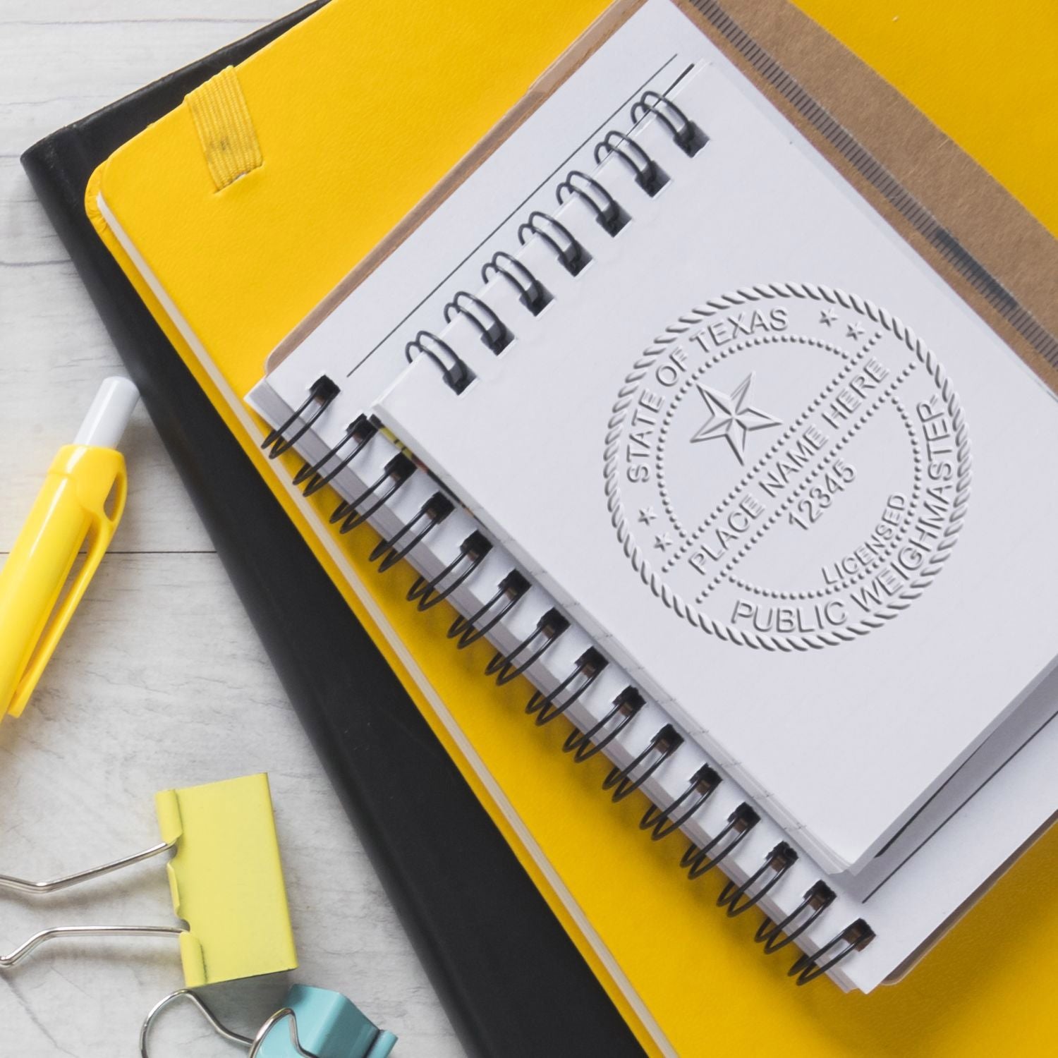 Yellow office stationery with spiral notebook featuring the Texas state seal, binder clips, and a pen on a desk.