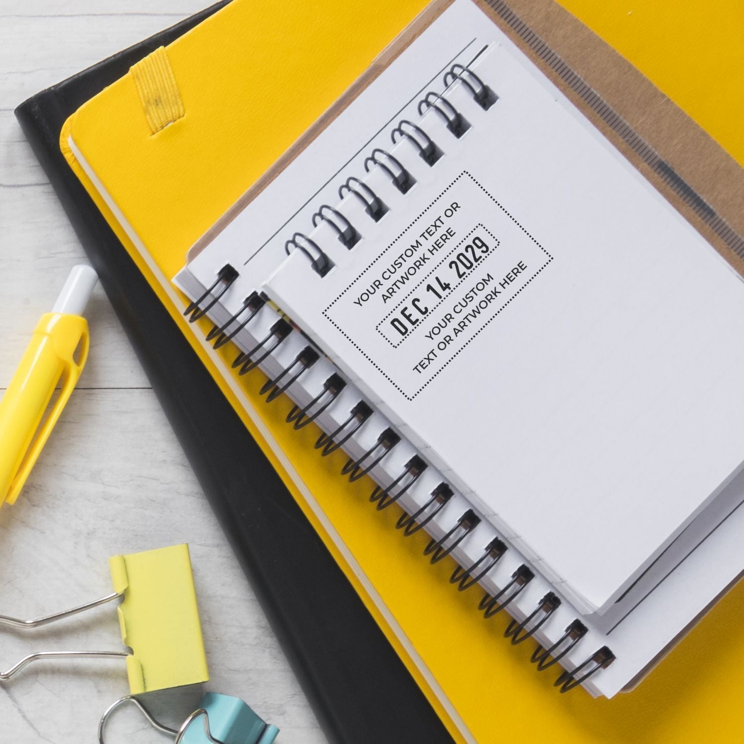 Top view of a yellow spiral notebook planner with a dated cover, yellow pen, and binder clips on a white desk, office stationery flat lay.