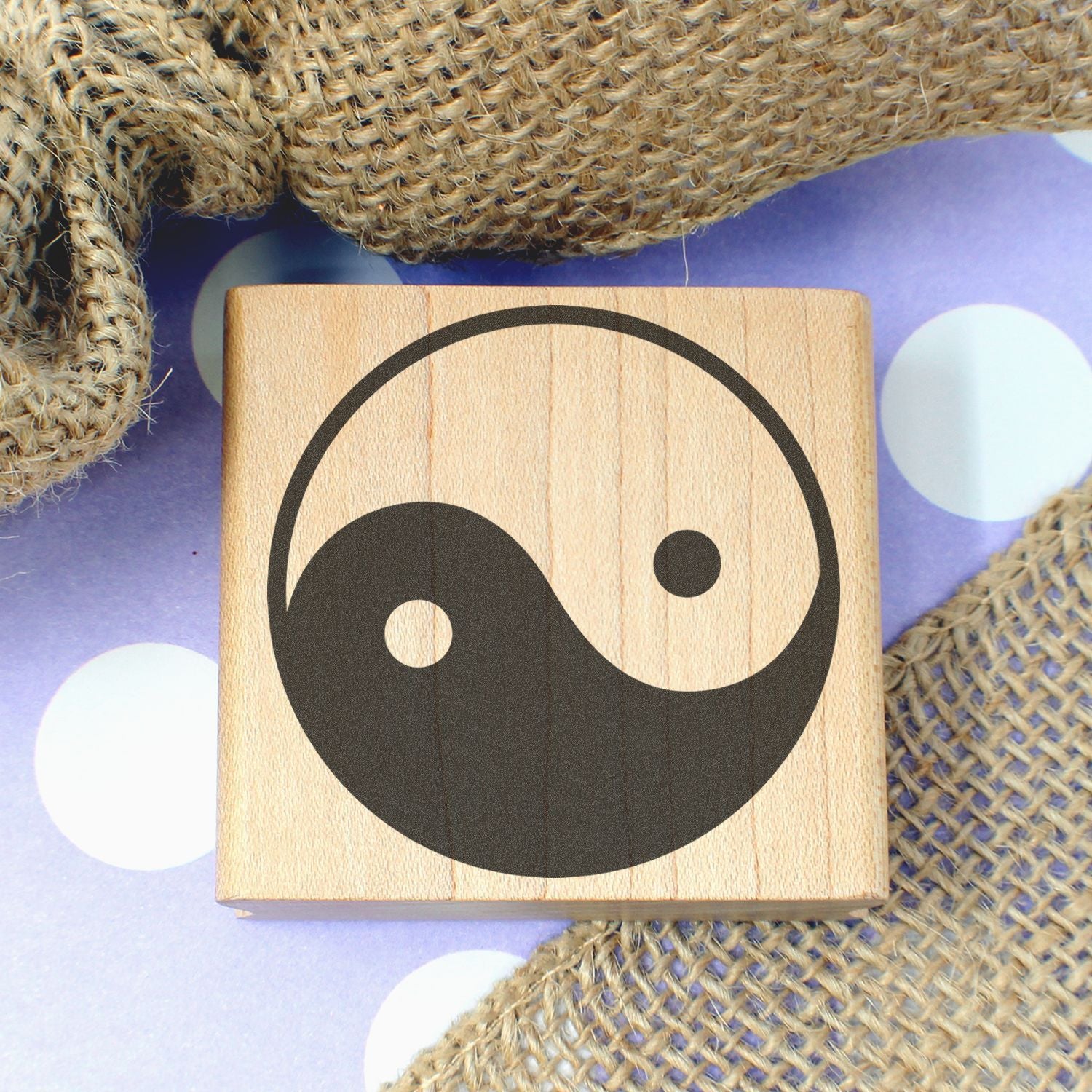 Wooden Yin Yang symbol block on burlap with purple polka dot background, minimalist spiritual decor.